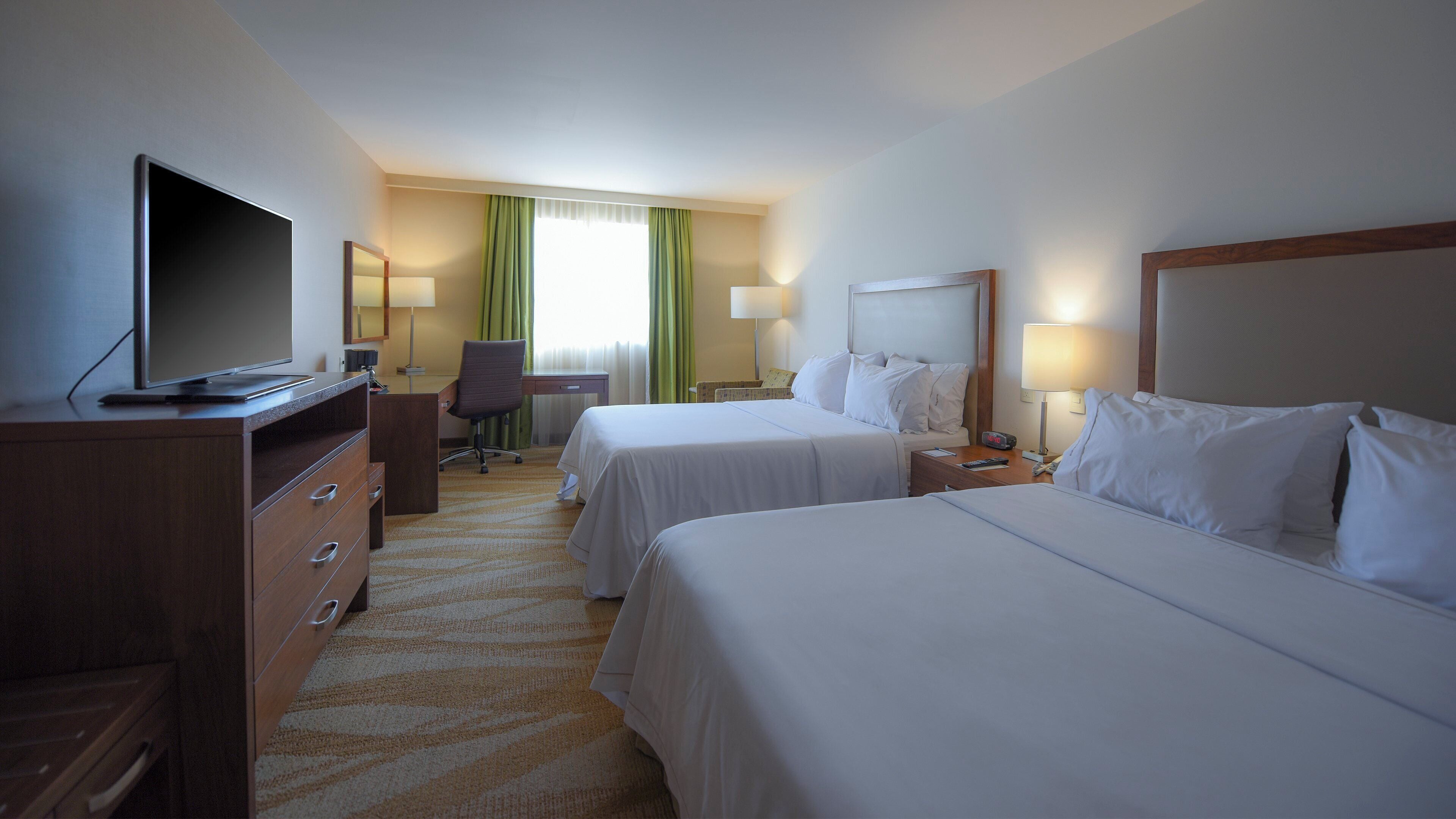 A hotel room with two beds, a desk, a chair, a television, and a lamp.