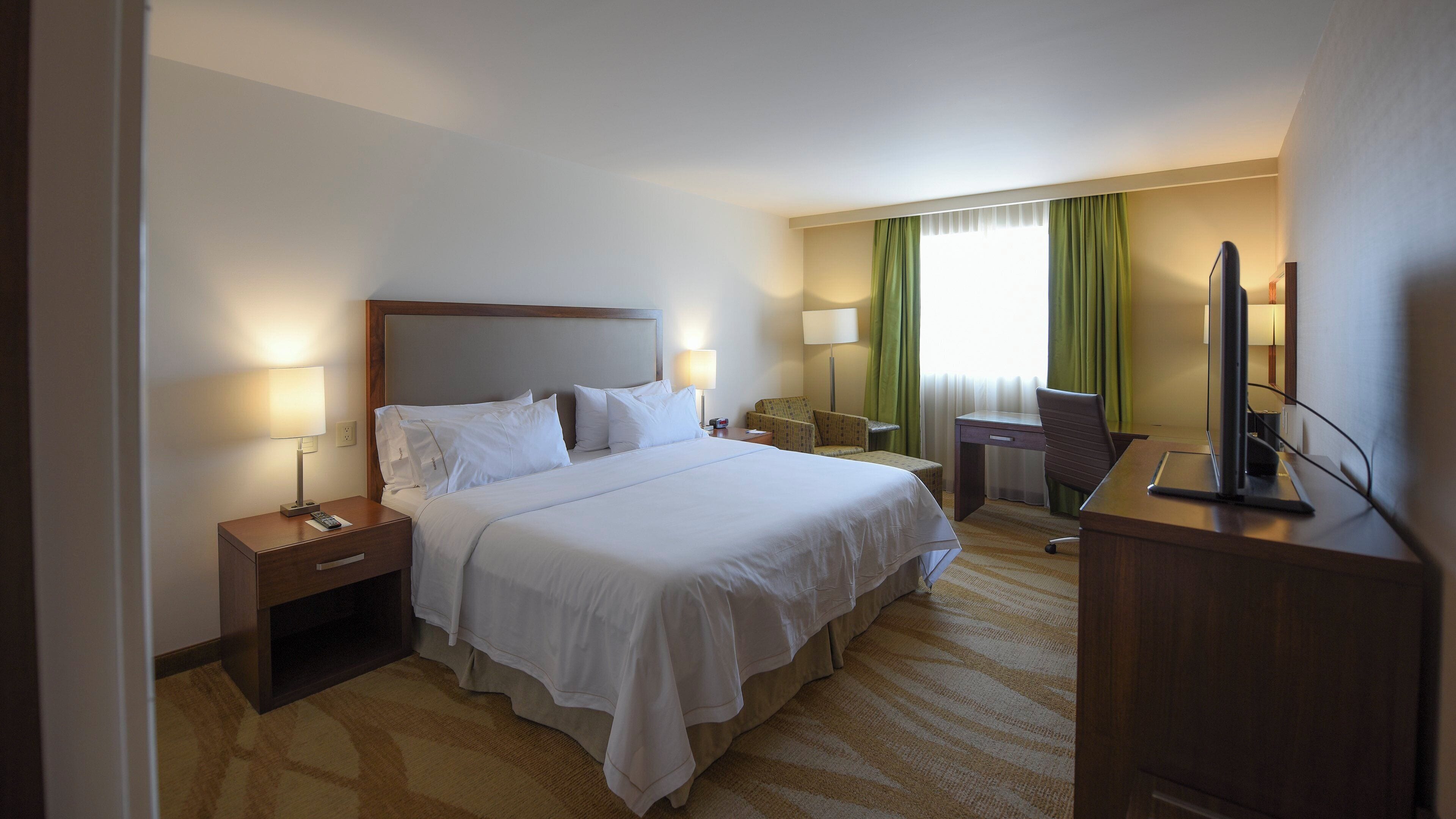 A hotel room with a large bed, bedside tables, a desk, a chair, and a television.