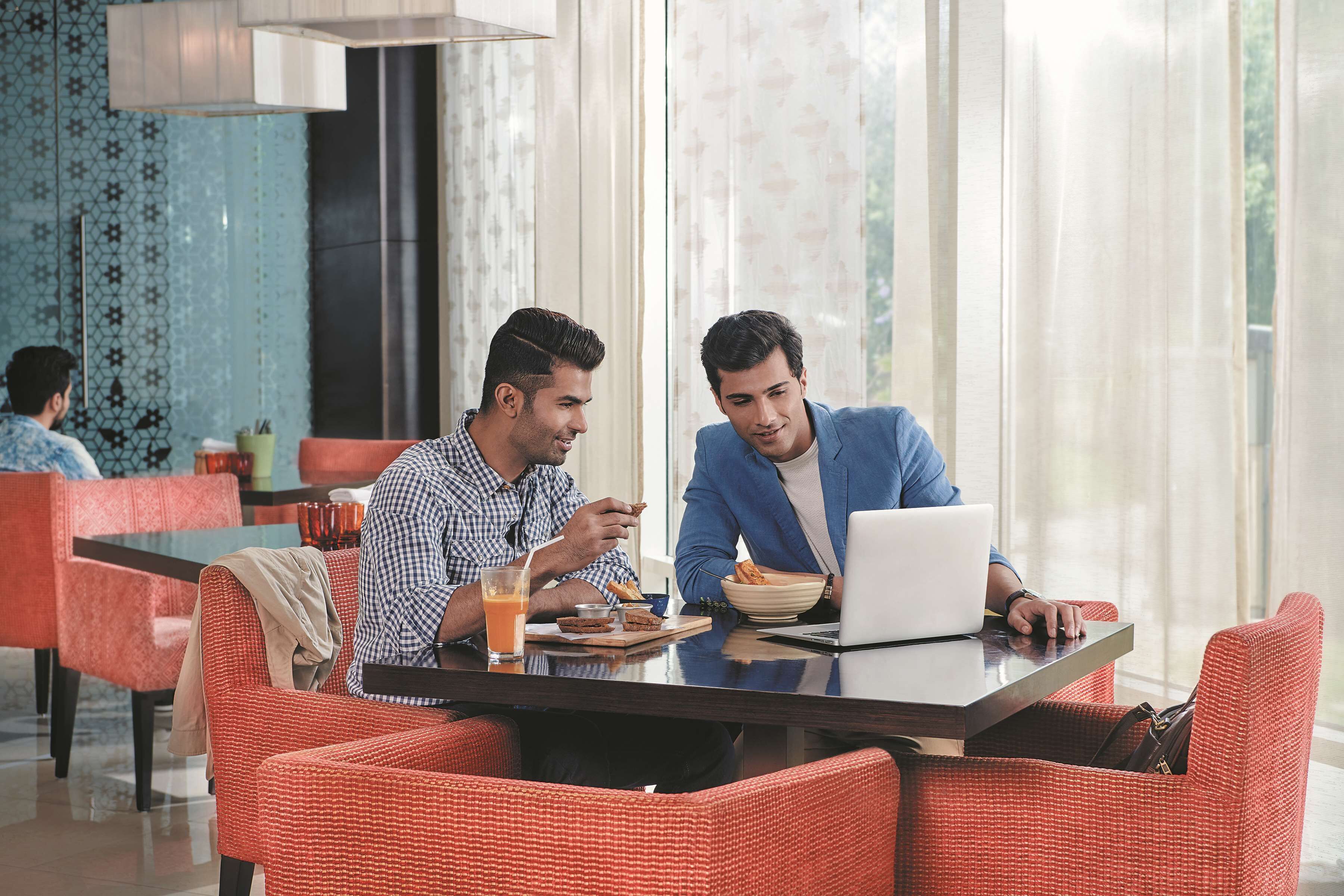 Two men sitting at a table with a laptop, orange juice, and food. The room has red chairs and a large window.