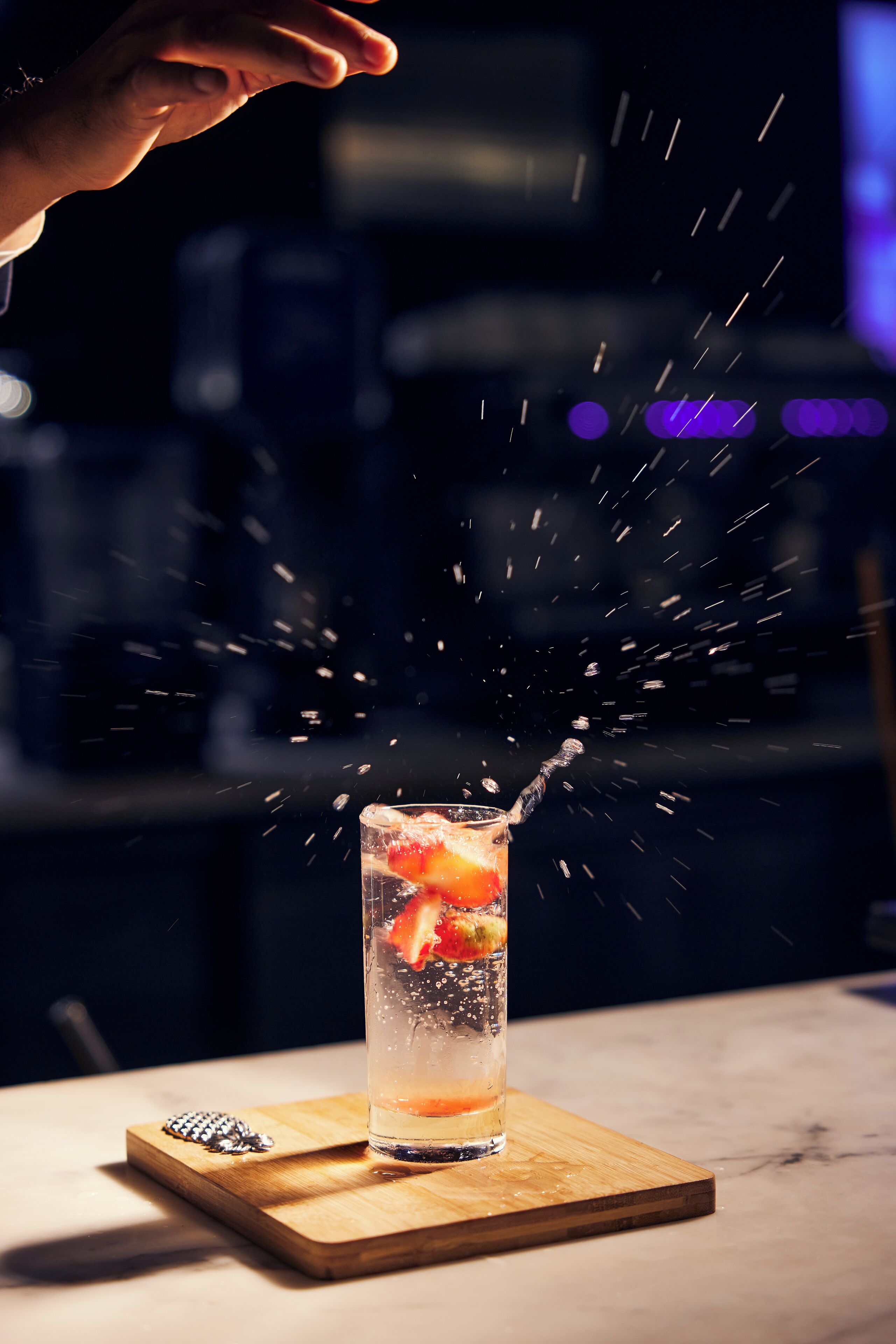 A tall glass with a drink, ice, and fruit on a wooden board.