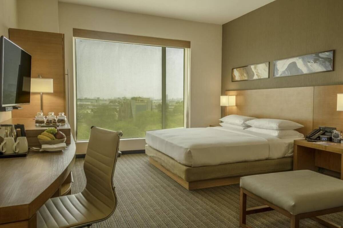A hotel room with a bed, a desk with a TV, a chair, a bedside table with a lamp, and a window with a city view.