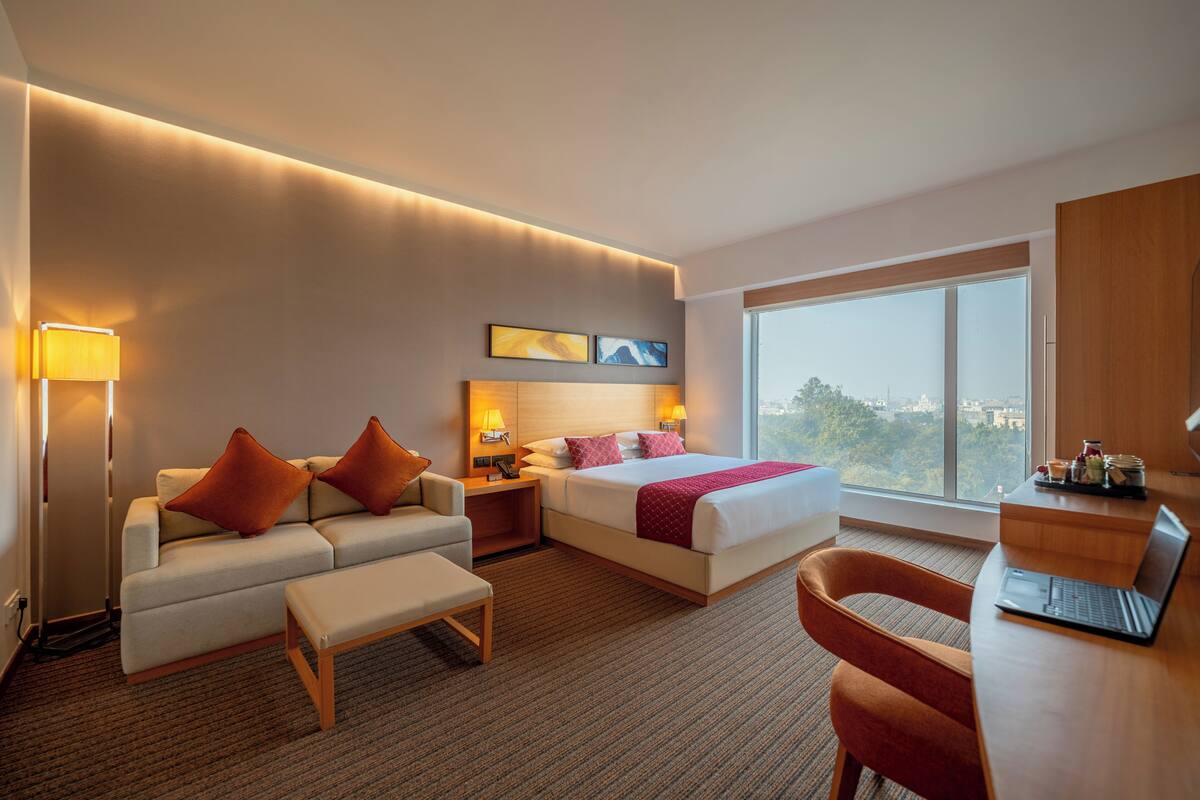 A hotel room with a bed, a sofa, a desk, and a large window offering a city view.