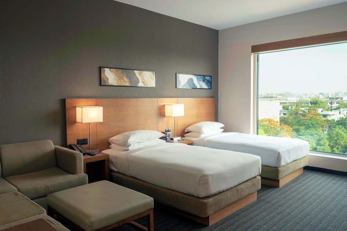 A hotel room with two beds, a sofa, a desk, and a large window offering a city view.