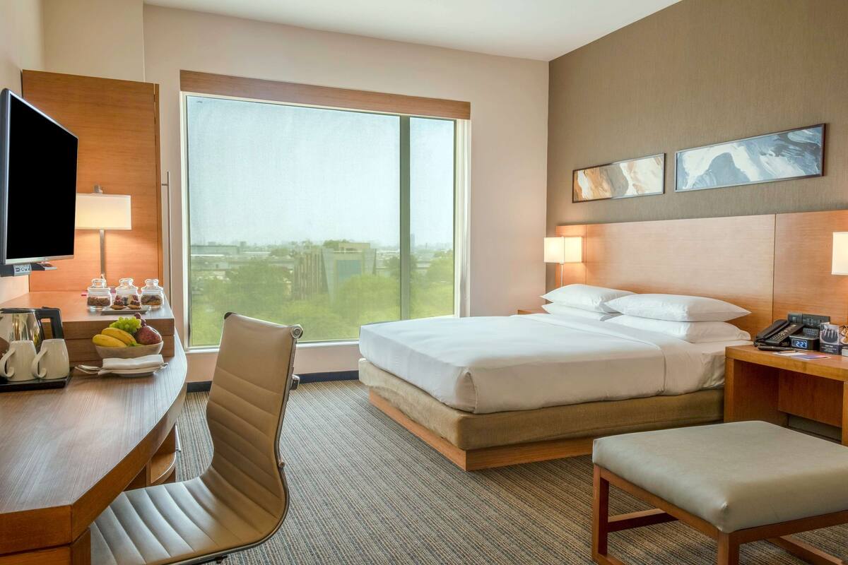 A hotel room with a bed, a desk with a television, a chair, and a view of a cityscape through large windows.
