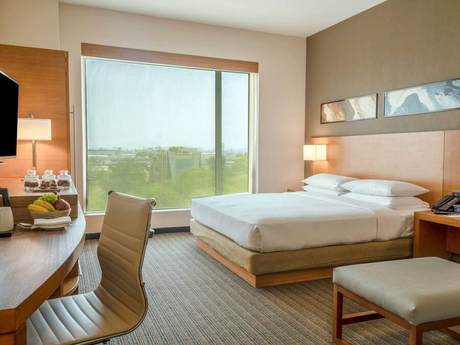 Ein Hotelzimmer mit Bett, Schreibtisch, Fernseher, Stuhl und Blick auf eine Stadtkulisse durch große Fenster.