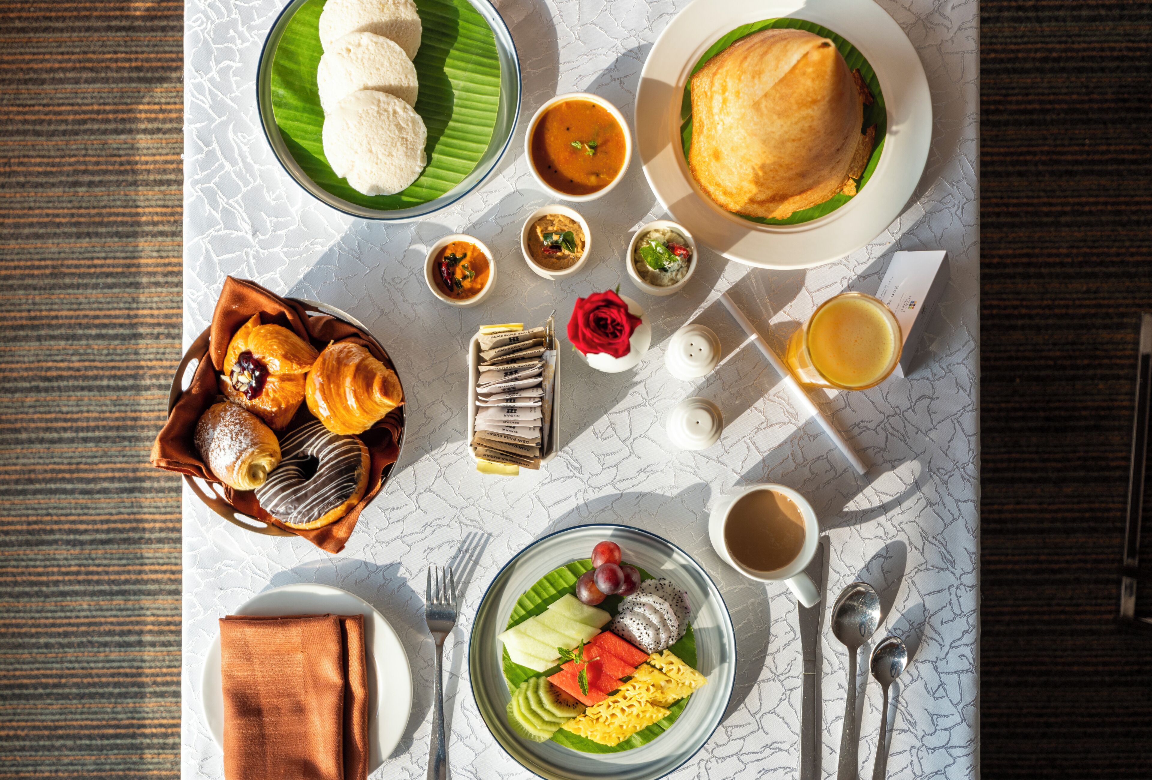 Une table dressée avec divers plats, notamment du pain, de la soupe et des fruits, ainsi qu’une rose et un verre de jus.