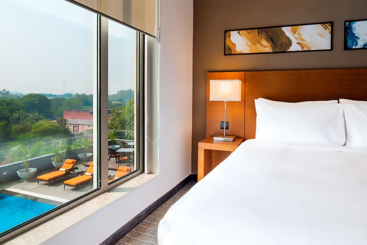 A hotel room with a large bed, a nightstand with a lamp, a view of a swimming pool, and two artworks on the wall.