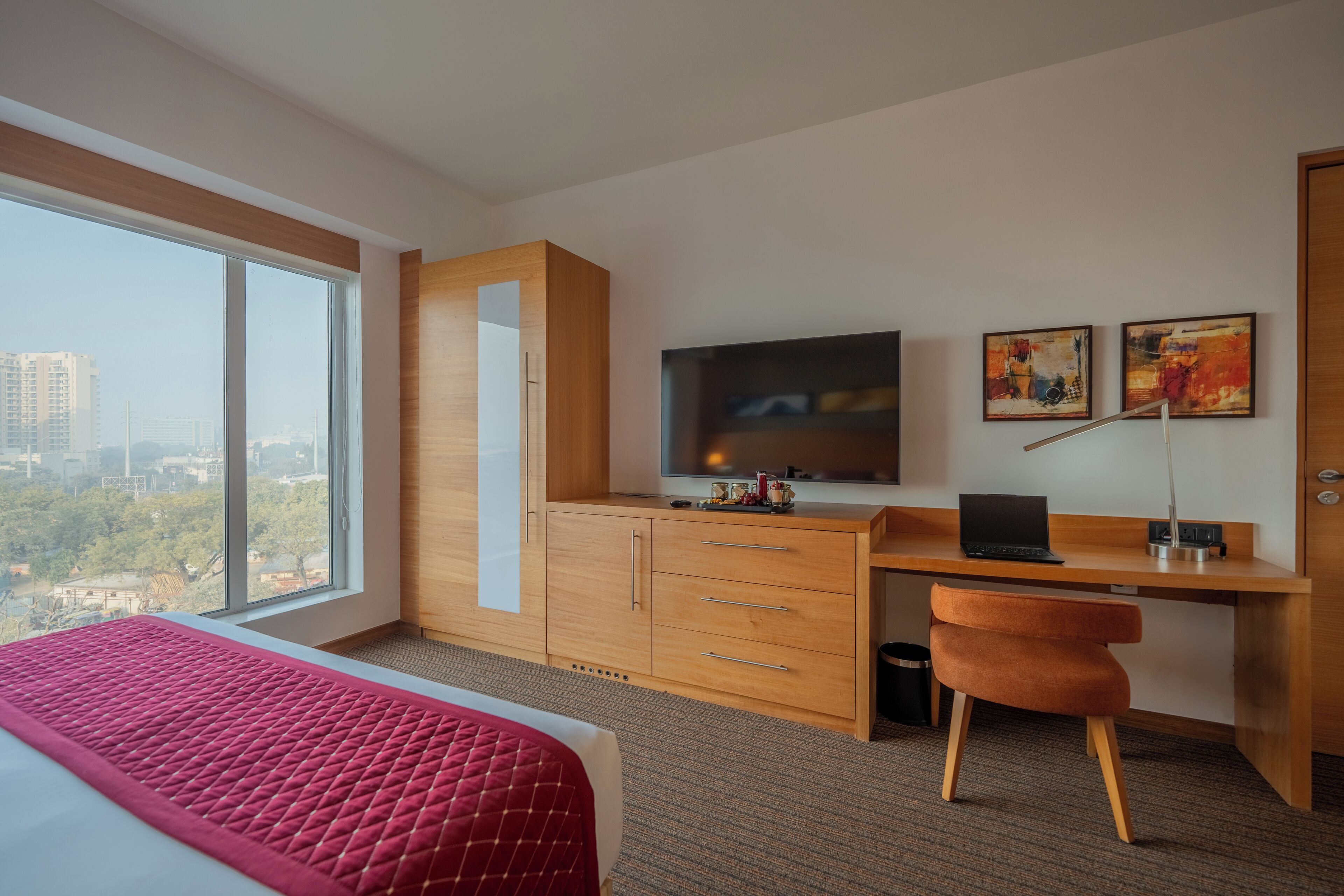 A hotel room with a large window, a bed with a red quilt, a wooden desk with a laptop, a TV, and two framed artworks on the wall.