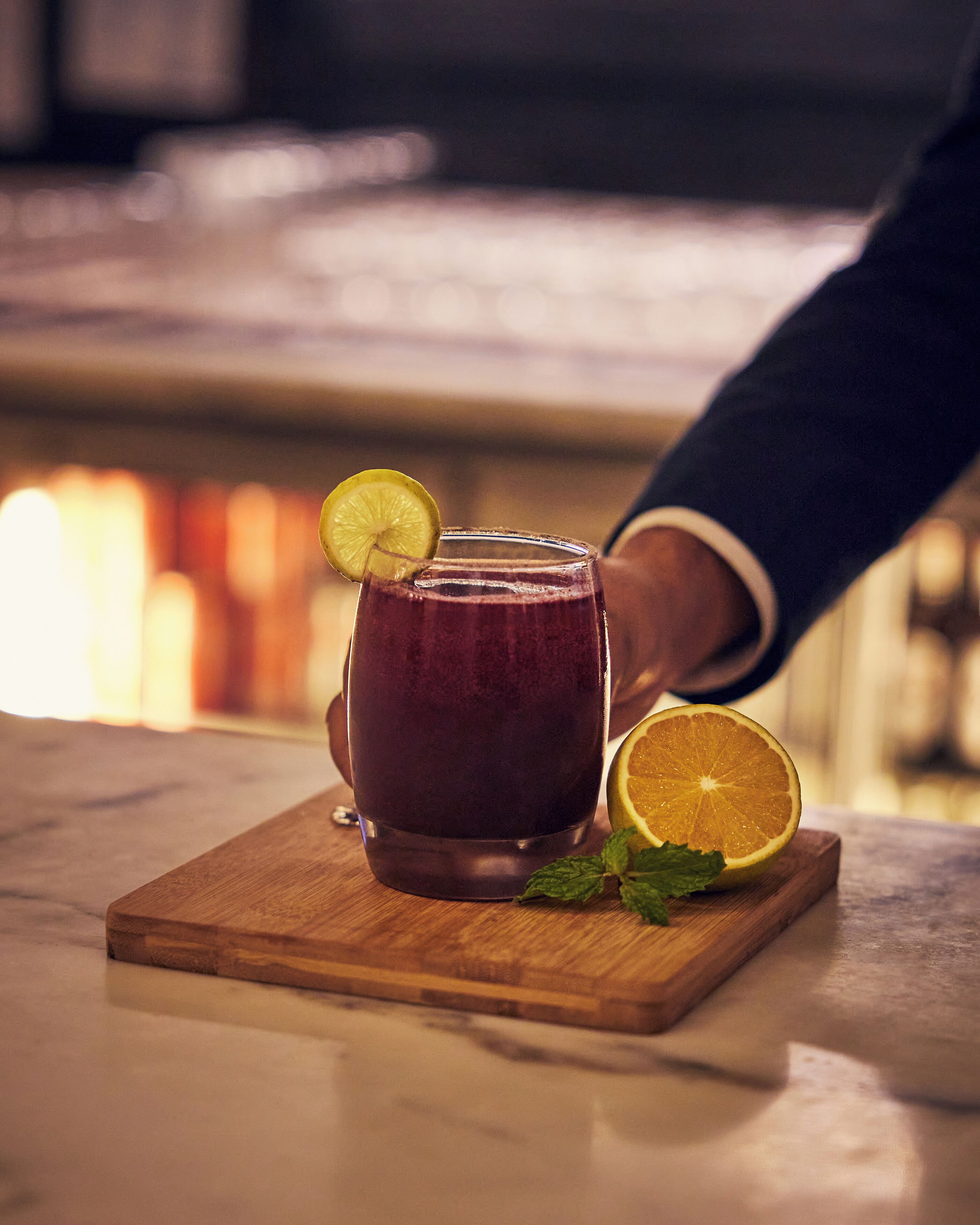 A glass of red beverage with a lime slice garnish on a wooden board, accompanied by a half orange and a mint leaf.