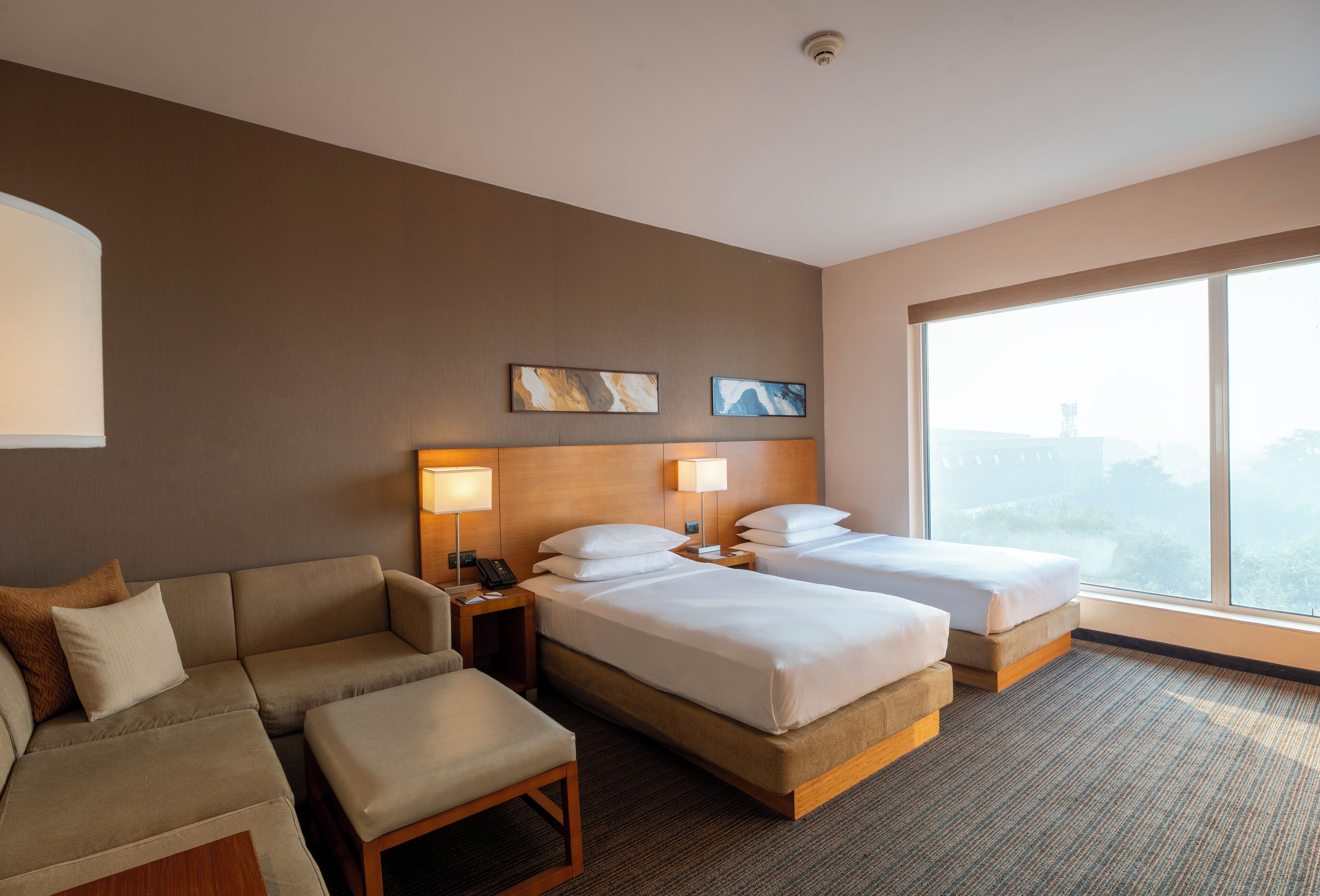 A hotel room with a sofa, two beds, a nightstand, and a large window.