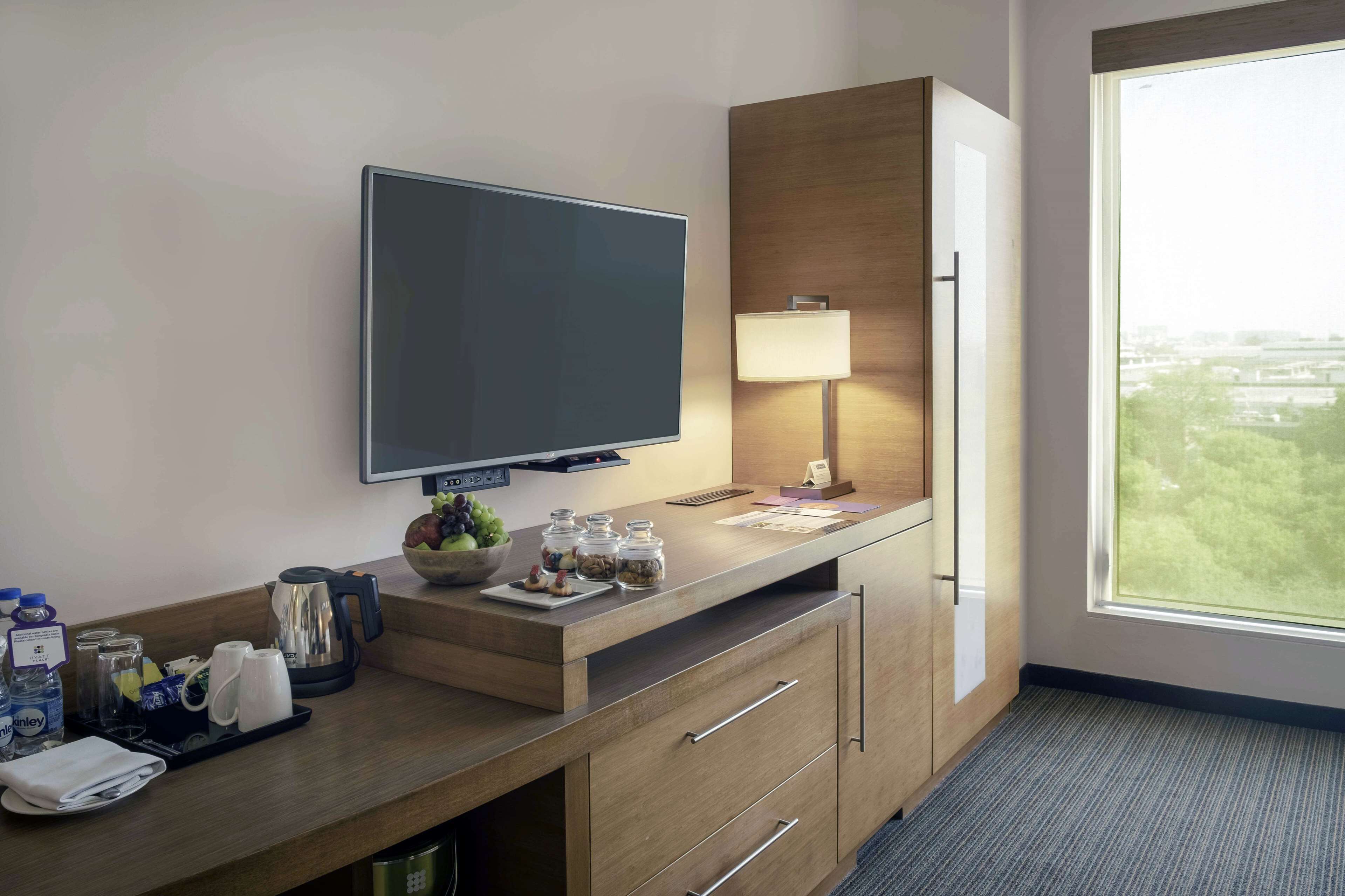 Hotelkamer met een flatscreen televisie, een houten bureau met lamp, een koffiezetapparaat en uitzicht op de stad door een groot raam.