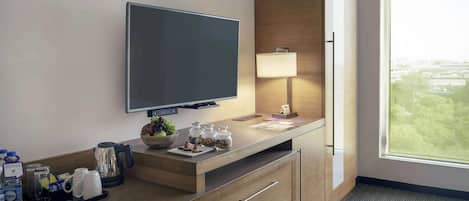 Habitación de hotel con televisor de pantalla plana, escritorio de madera con lámpara, cafetera y vistas a la ciudad a través de un ventanal grande.