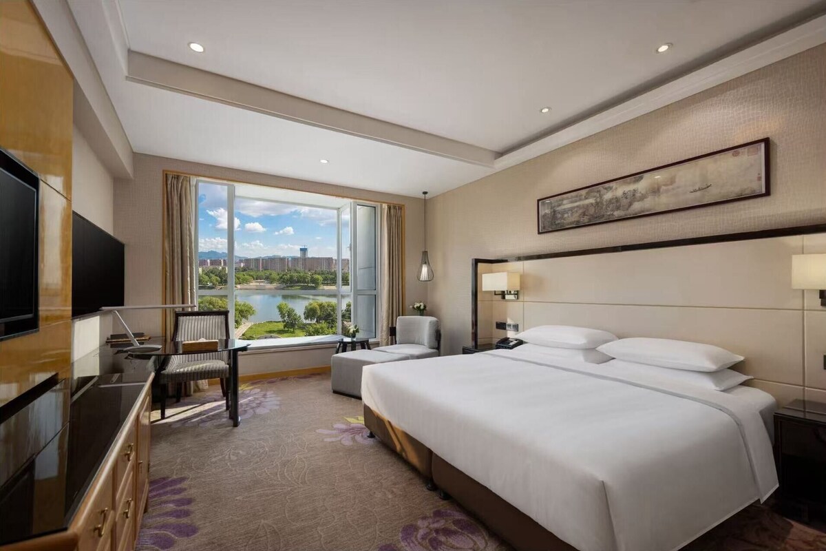 Quarto de hotel com uma cama grande, uma escrivaninha com cadeira, televisão e vista para a paisagem urbana e rio.