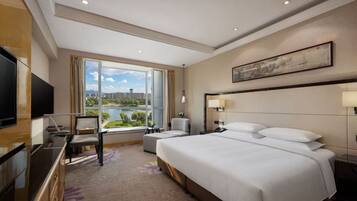 A hotel room with a large bed, a desk with a chair, a TV, and a view of a cityscape and river.