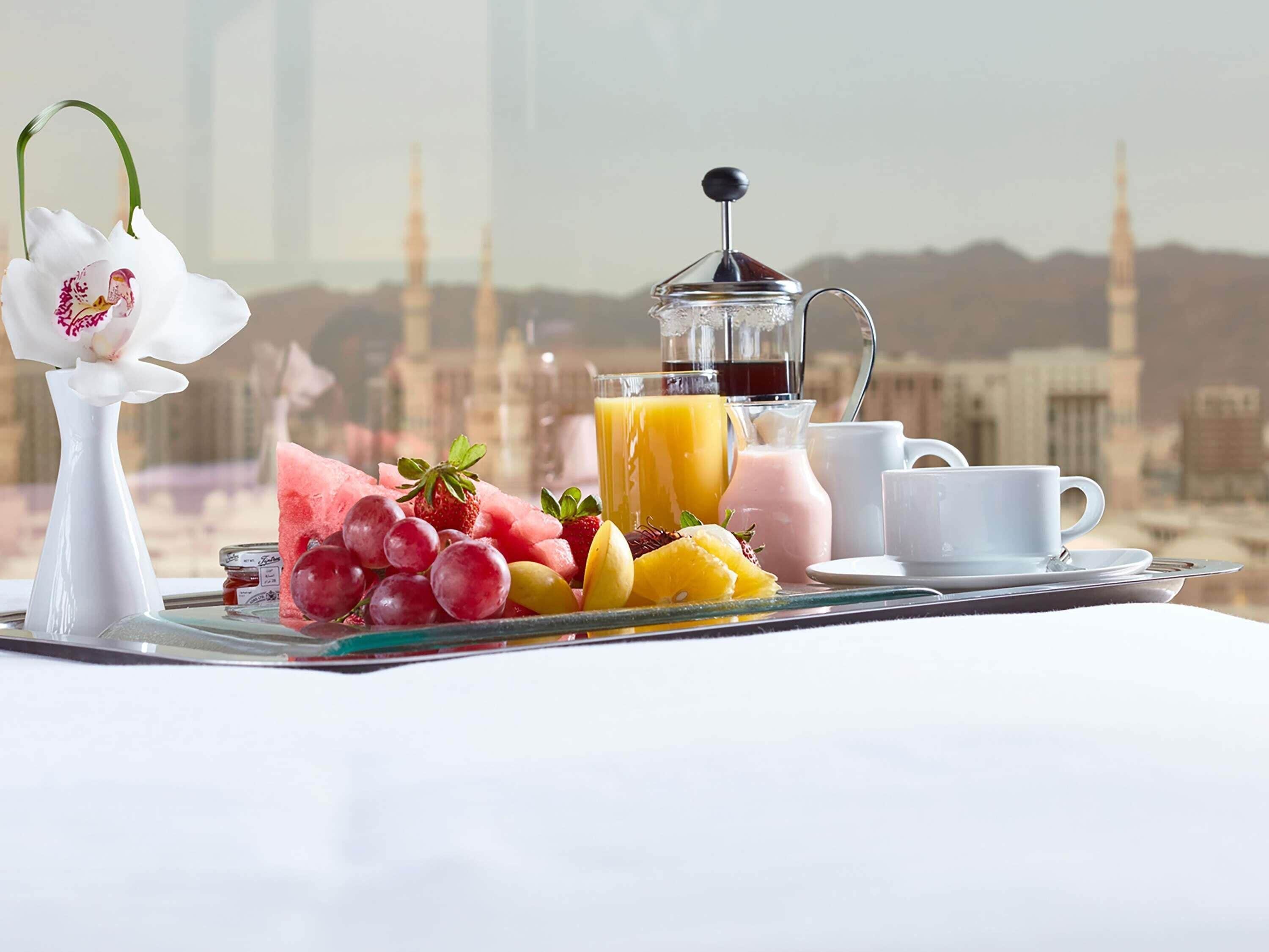 A breakfast tray with fruits, juices, and a coffee pot, set against a cityscape with minarets.