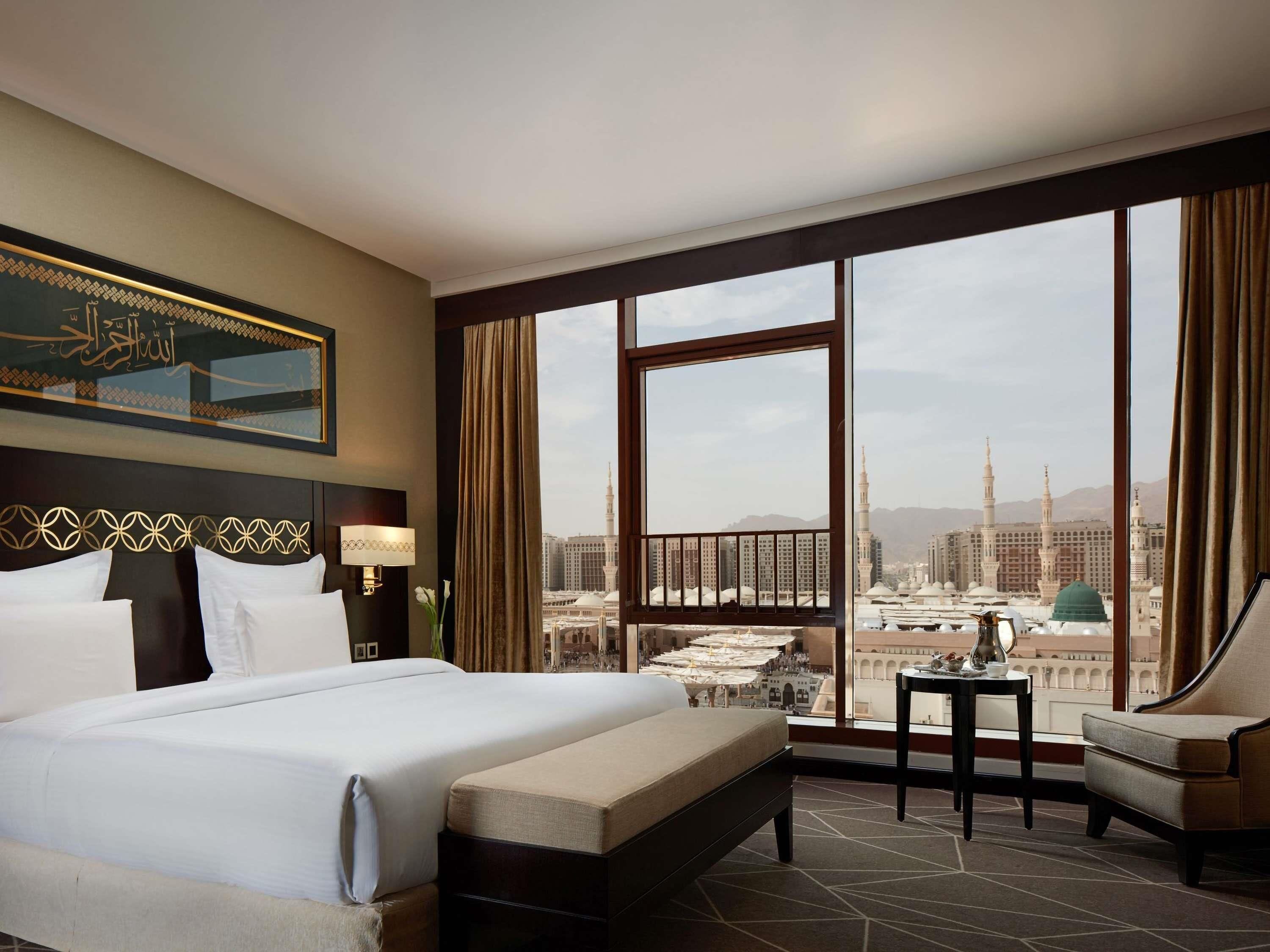 A hotel room with a large bed, a sofa, a small table, and a view of a cityscape with tall buildings and a mosque.
