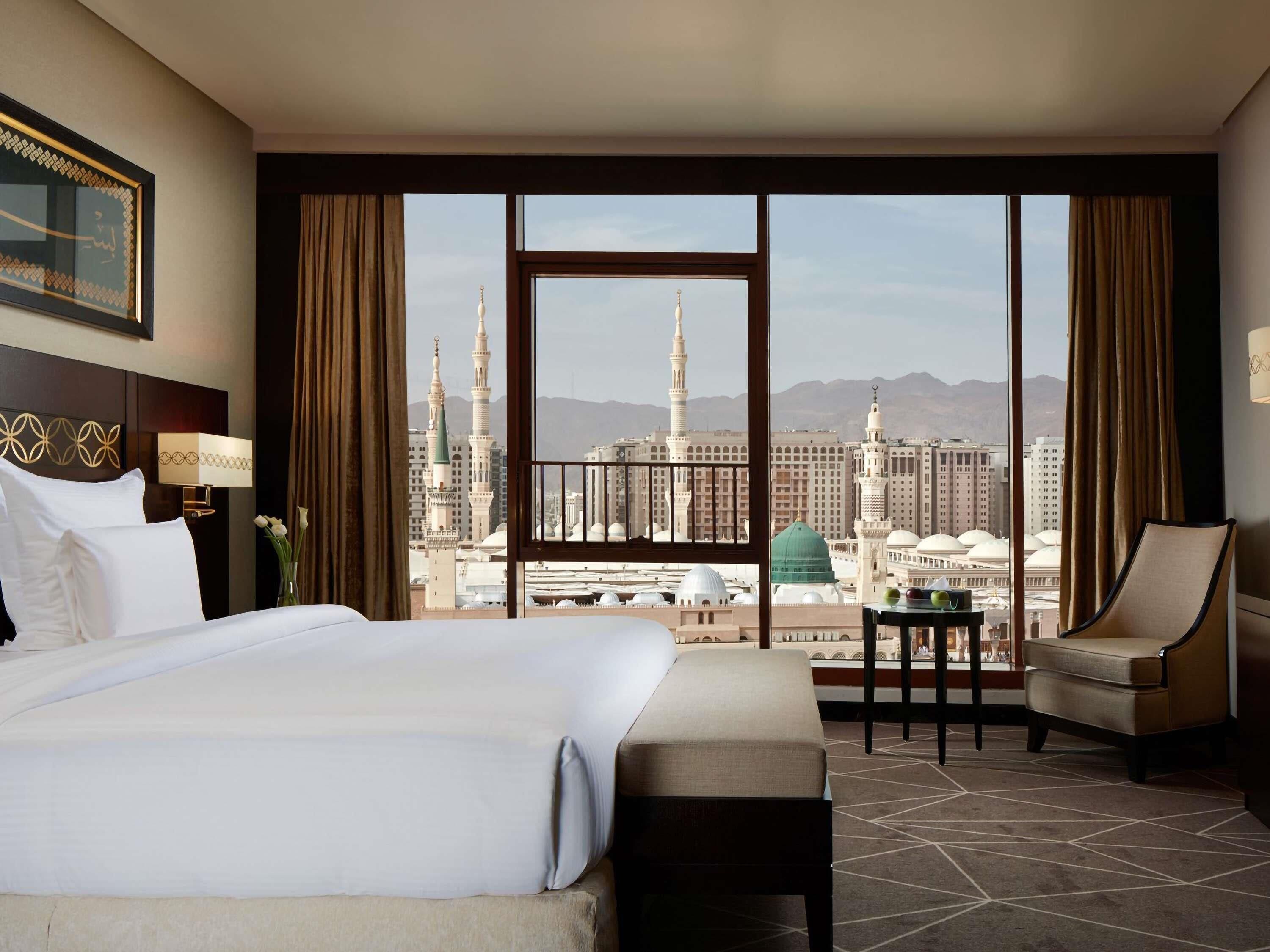 A hotel room with a large bed, a desk, a chair, and a view of a cityscape with prominent buildings and a mosque.
