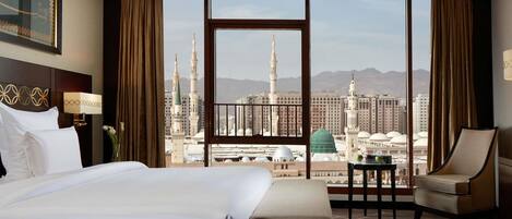 A hotel room with a large bed, a desk, a chair, and a view of a cityscape with prominent buildings and a mosque.