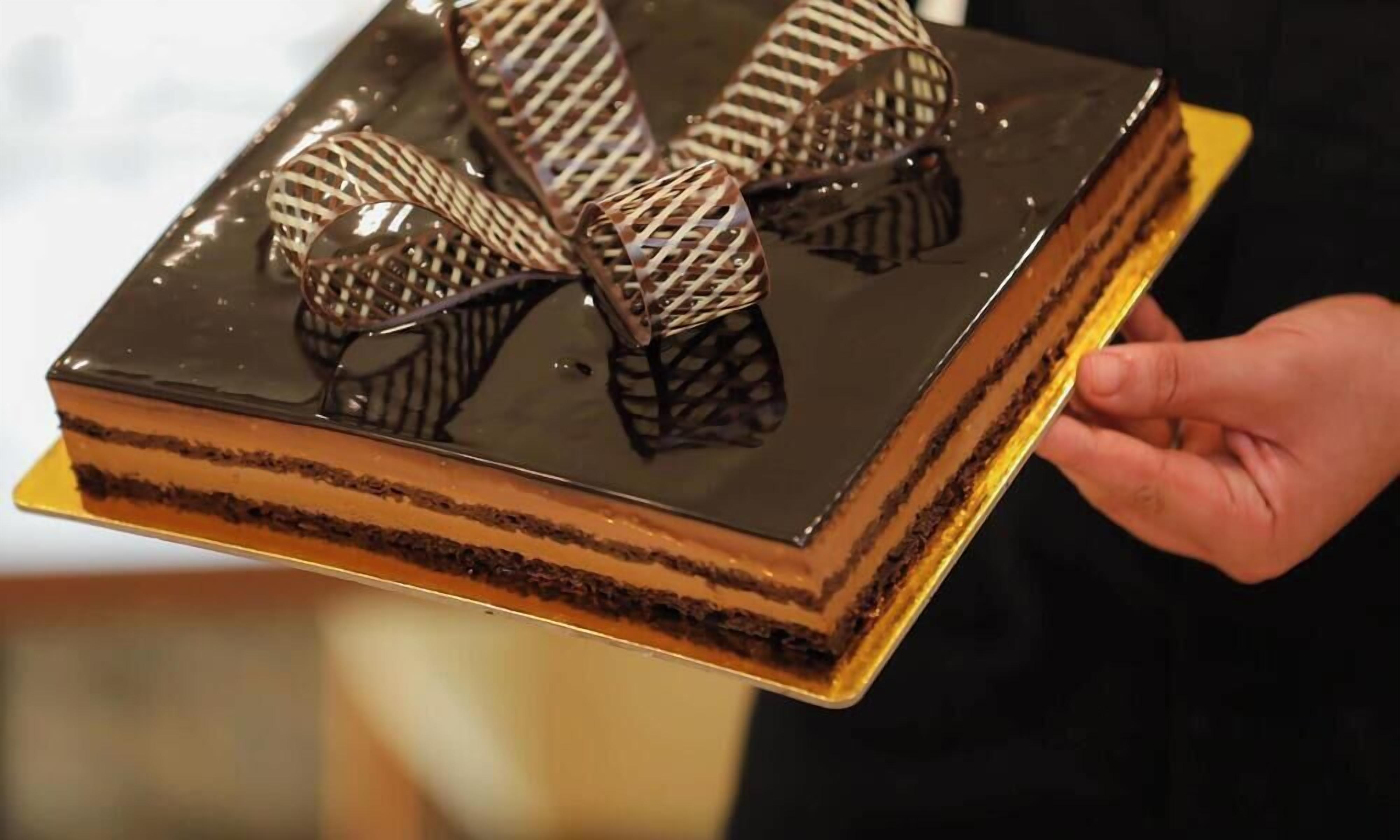 En person håller i en rektangulär chokladtårta med flera lager och dekorativa chokladbitar ovanpå.