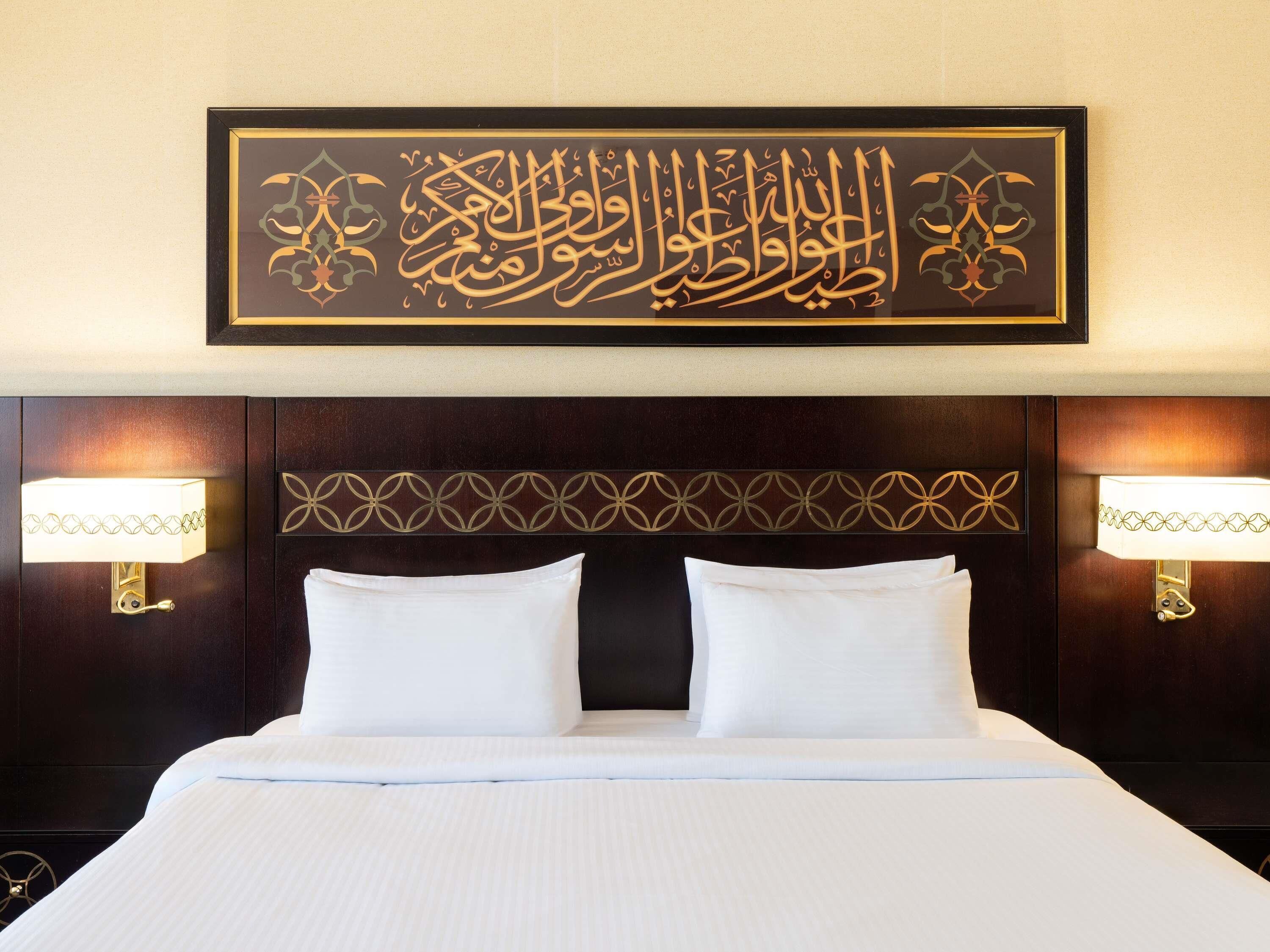 Una habitación de hotel con una cama grande, dos lámparas de noche y una obra de caligrafía árabe enmarcada en la pared.