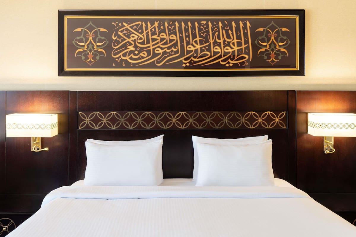 A hotel room with a large bed, two bedside lamps, and a framed Arabic calligraphy piece on the wall.