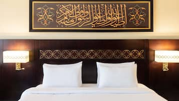 A hotel room with a large bed, two bedside lamps, and a framed Arabic calligraphy piece on the wall.