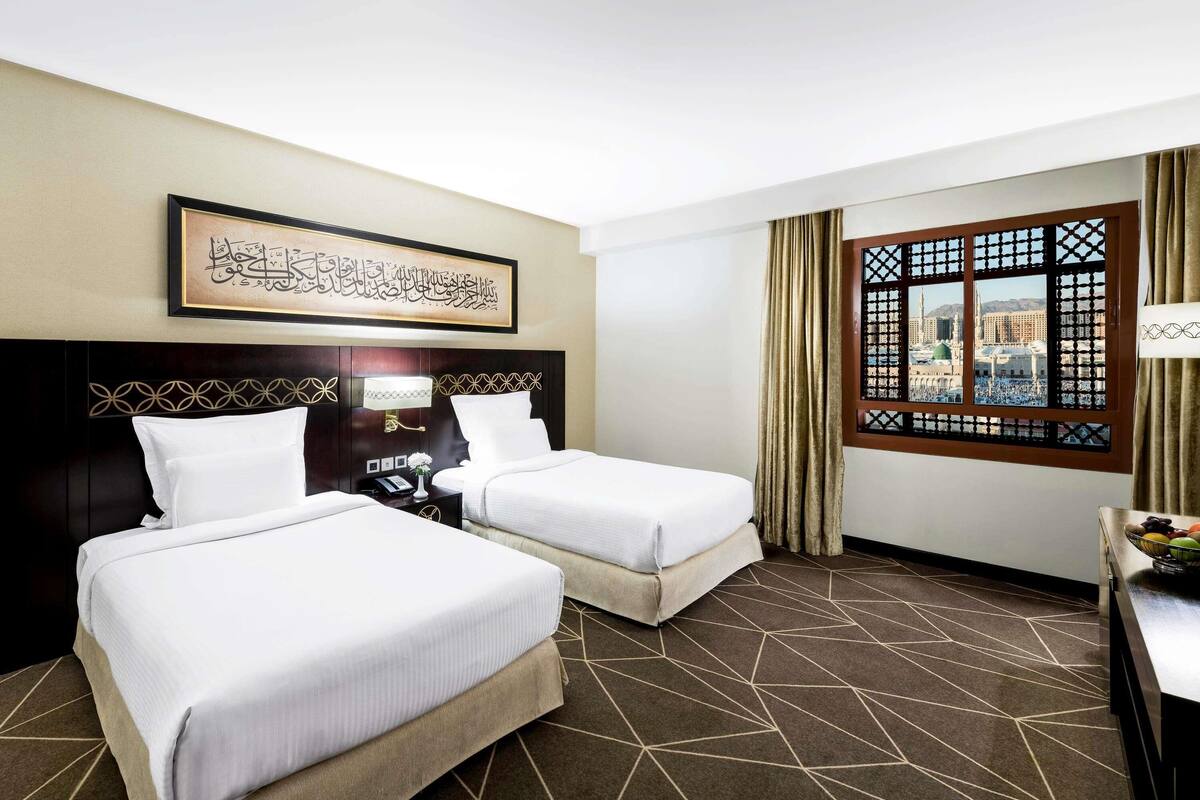 A hotel room with two beds, a nightstand, a framed calligraphy piece, a window with a view, and a patterned carpet.