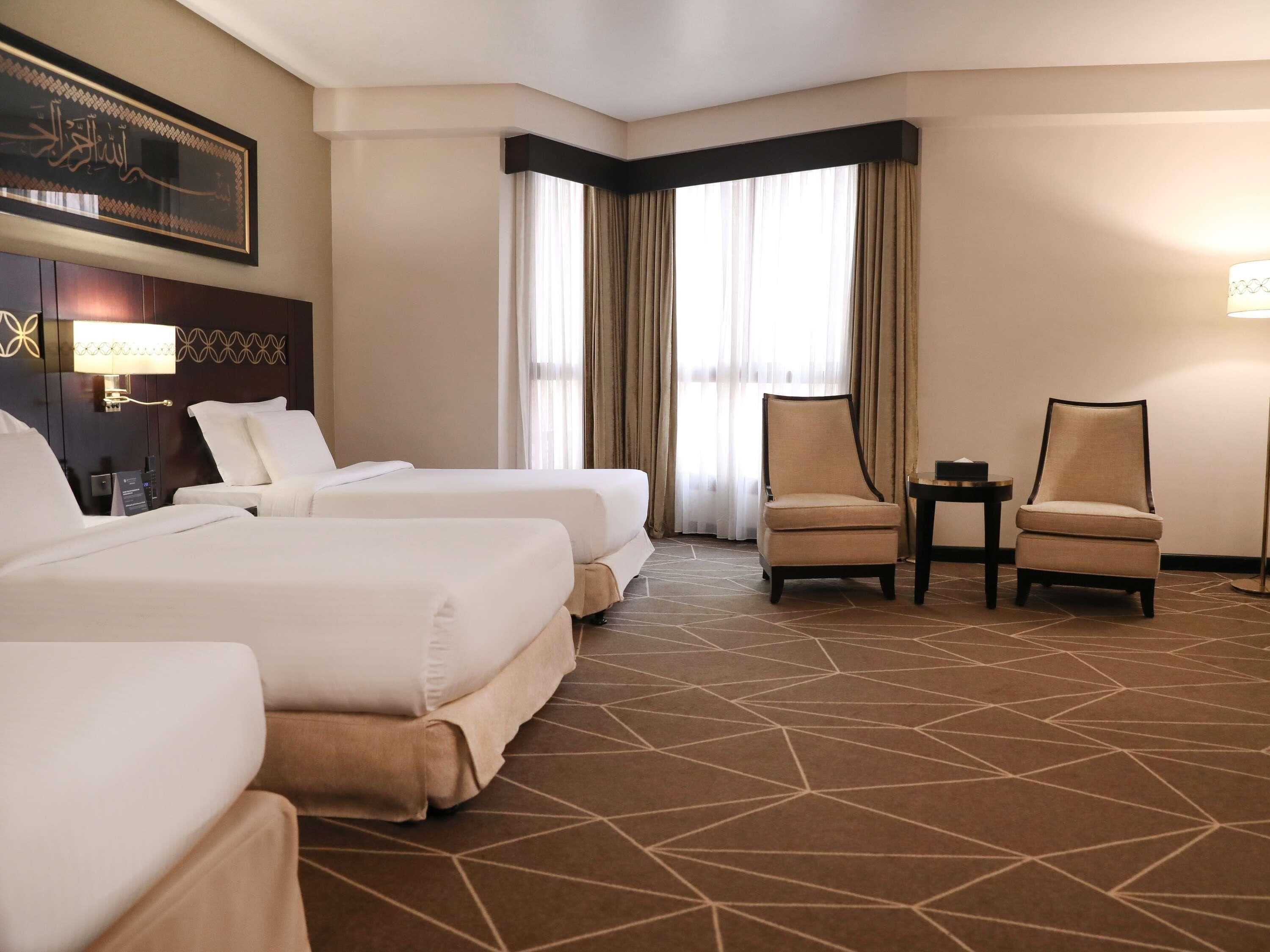 Habitación de hotel con dos camas, una silla, una mesita y una lámpara de pared.