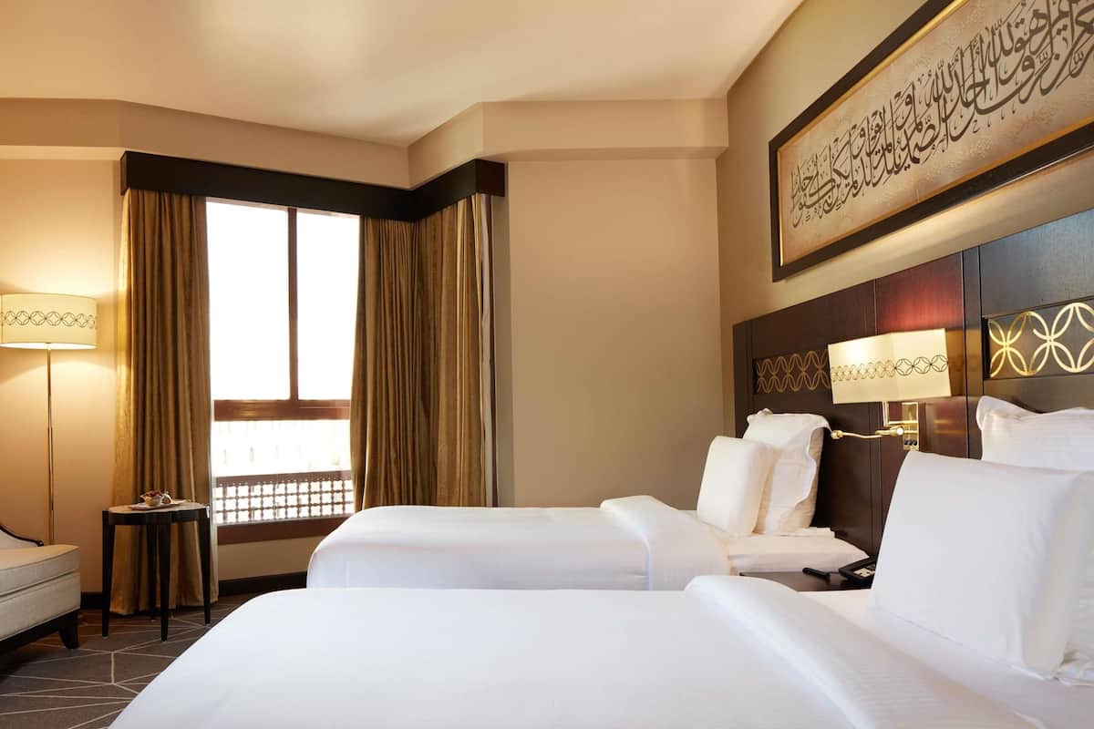 A hotel room with two beds, a bedside table, a lamp, a window with curtains, and a decorative wall piece.