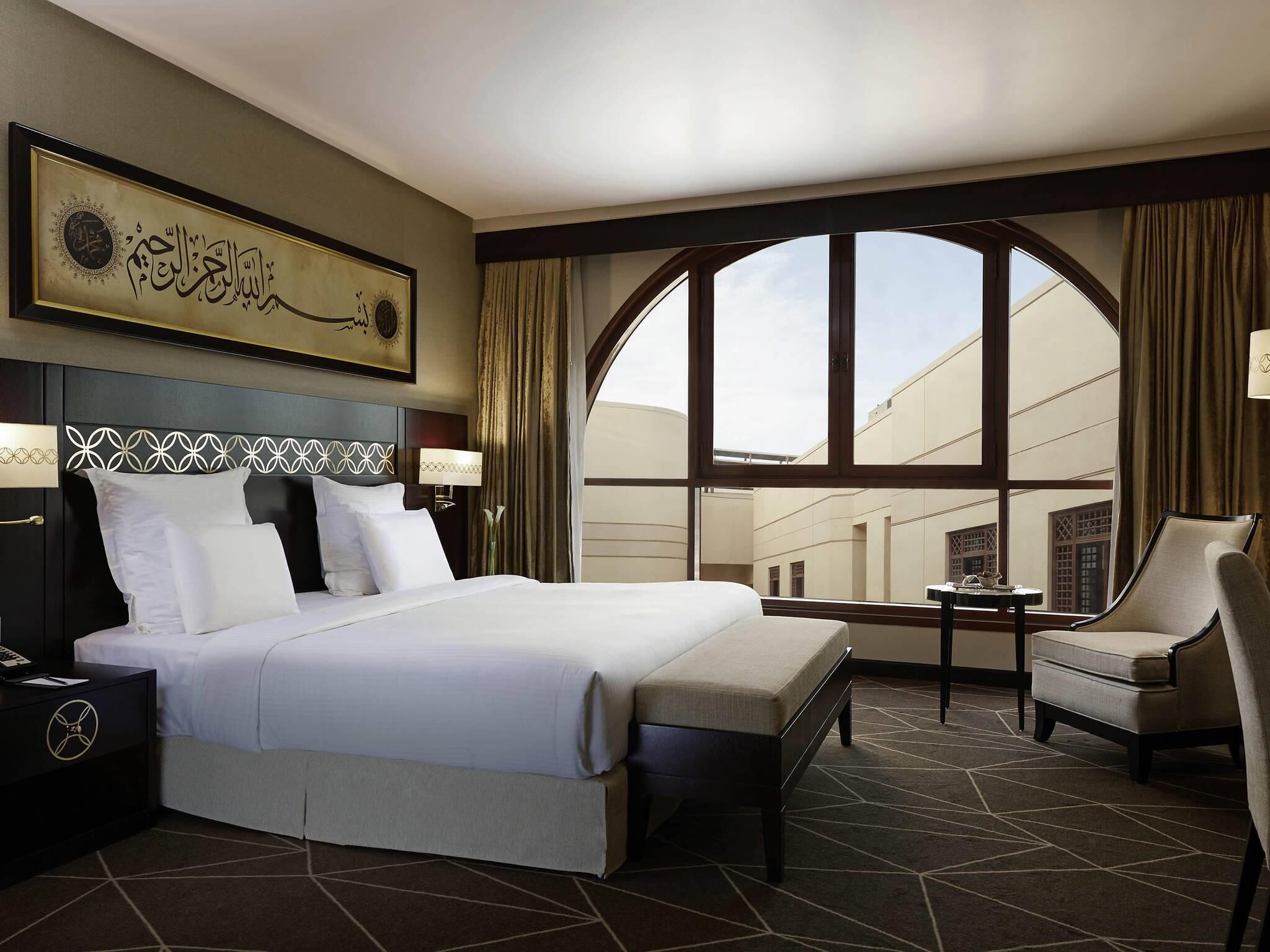 Une chambre d’hôtel avec un grand lit, un bureau, une chaise et une vue sur le paysage urbain, visible à travers une grande fenêtre.