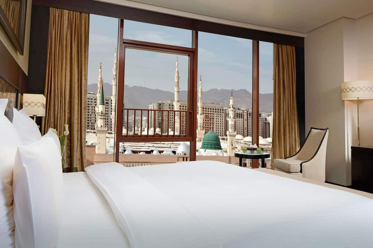 A hotel room with a large bed, a chair, a small table, and a view of a cityscape with buildings and mountains.