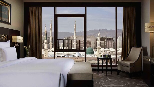 A hotel room with a large bed, a chair, a small table, and a view of a cityscape with tall buildings and a mosque.