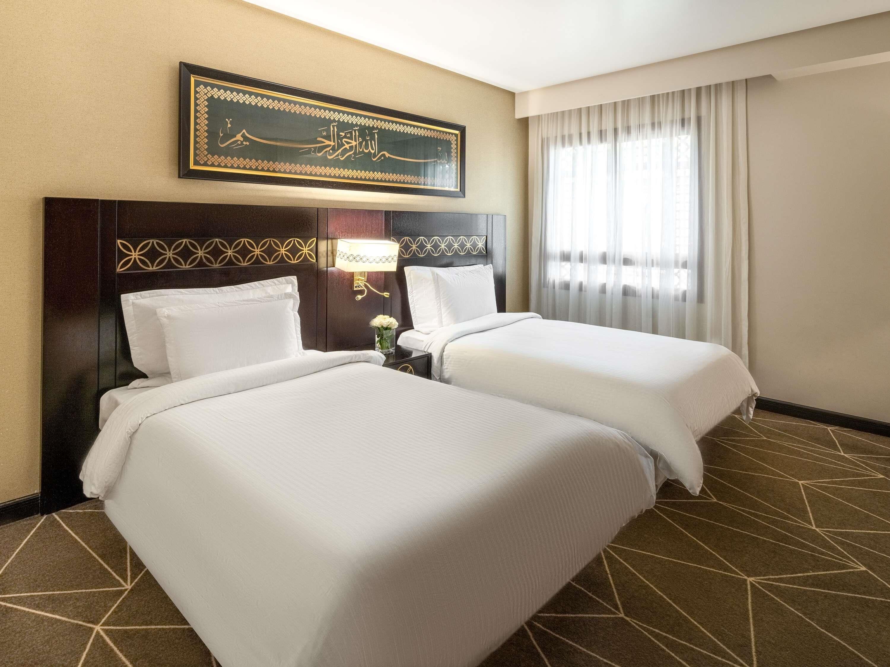 A hotel room with two beds, a decorative wall piece, a nightstand with a lamp, and a window with curtains.