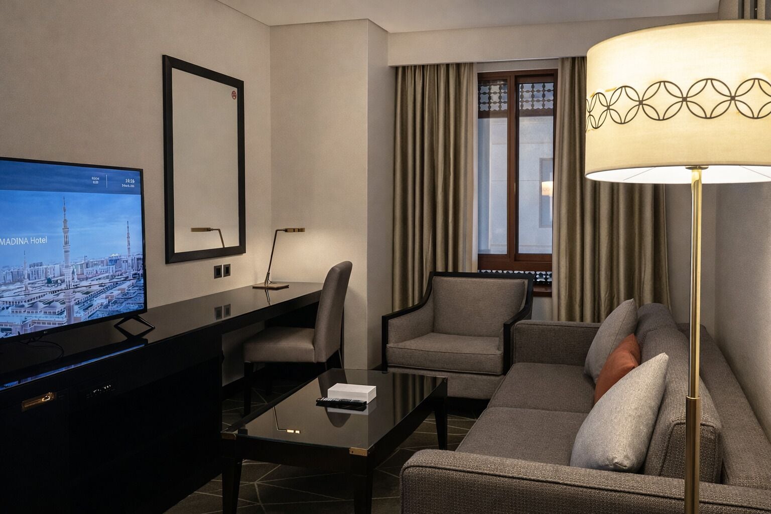A hotel room with a flat-screen TV displaying a cityscape, a desk with a lamp, two armchairs, a sofa, and a large floor lamp.