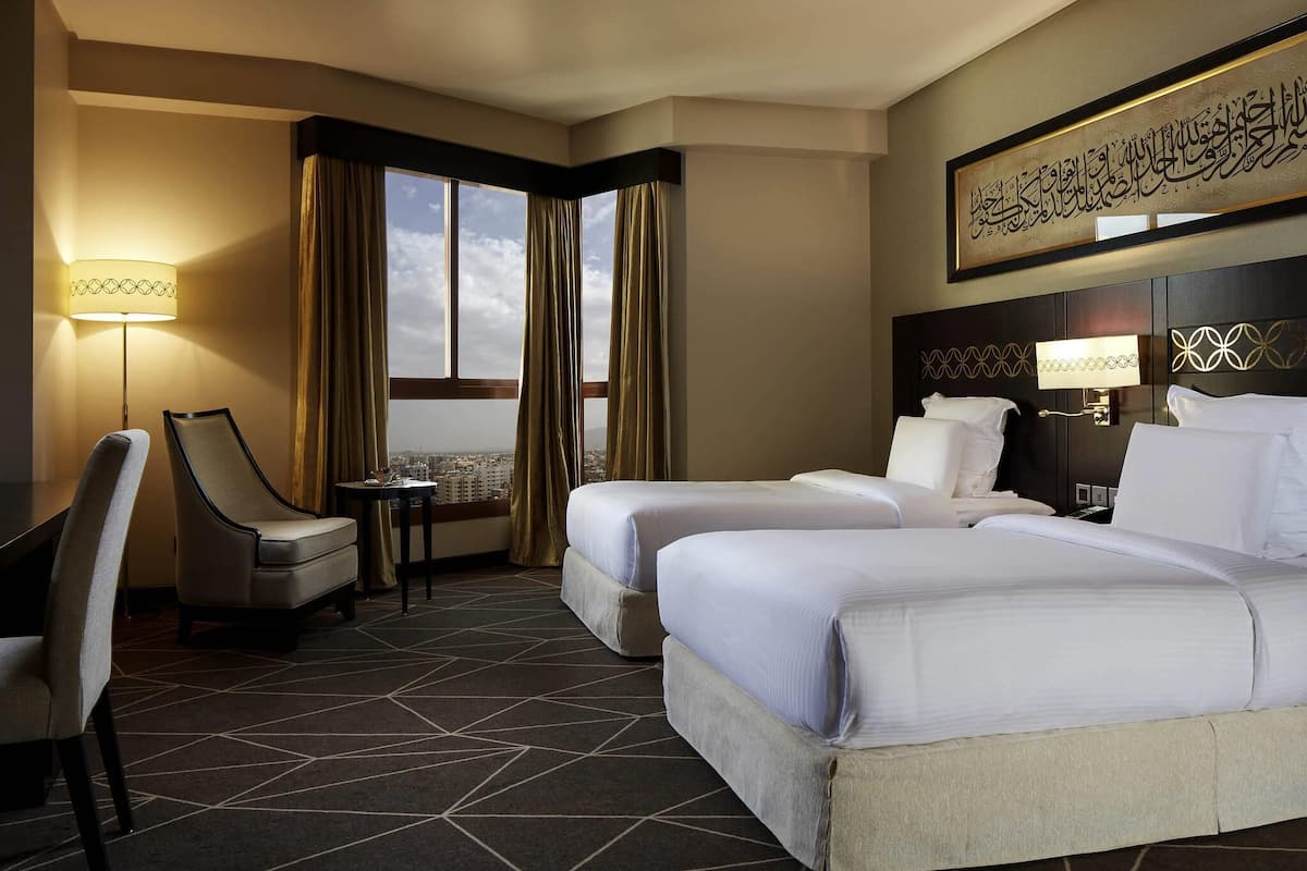 A hotel room with two beds, a desk, a chair, a lamp, and a view of the cityscape.