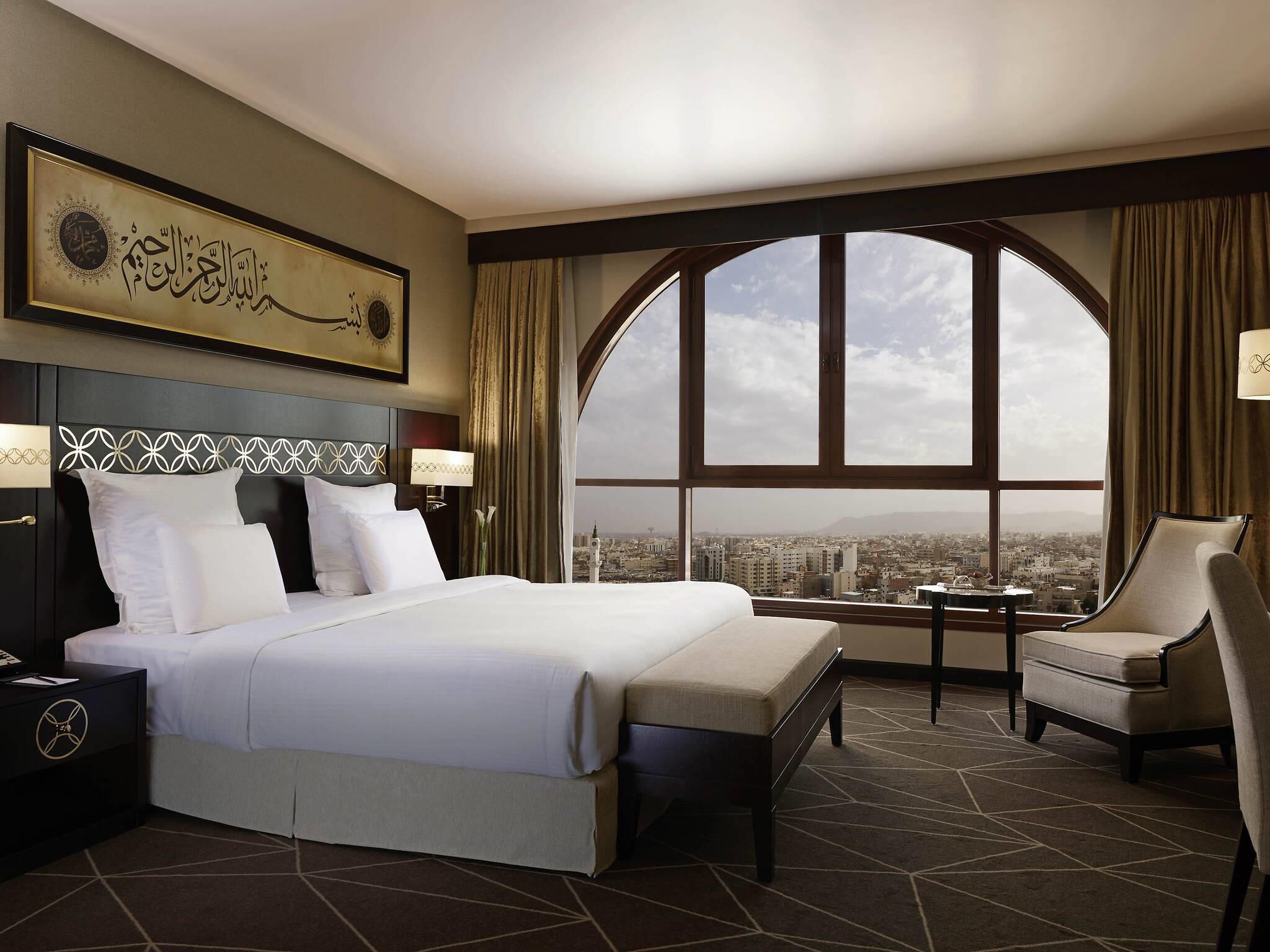 Une chambre d’hôtel avec un grand lit, une chaise, une table de chevet, une lampe, une vue sur le paysage urbain par une grande fenêtre et un élément décoratif mural.