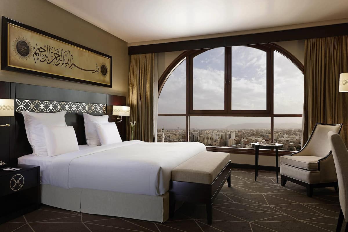 A hotel room with a large bed, a chair, a nightstand, a lamp, a view of a cityscape through a large window, and a decorative wall piece.