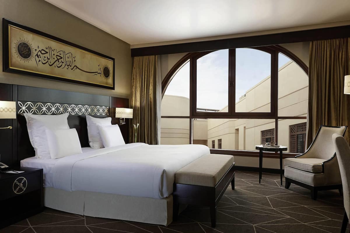 A hotel room with a large bed, a desk, a chair, and a view of the cityscape through a large window.