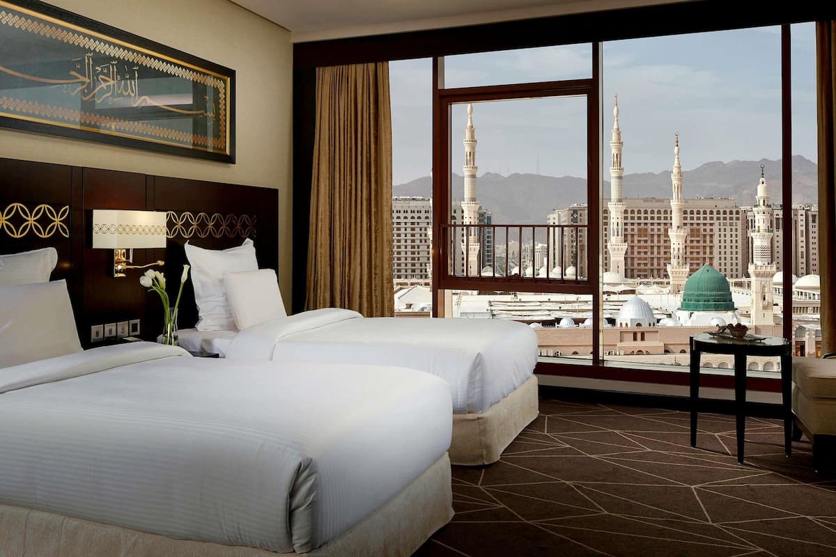 A hotel room with two beds, a large window with a view of a mosque and cityscape, and a decorative wall piece.