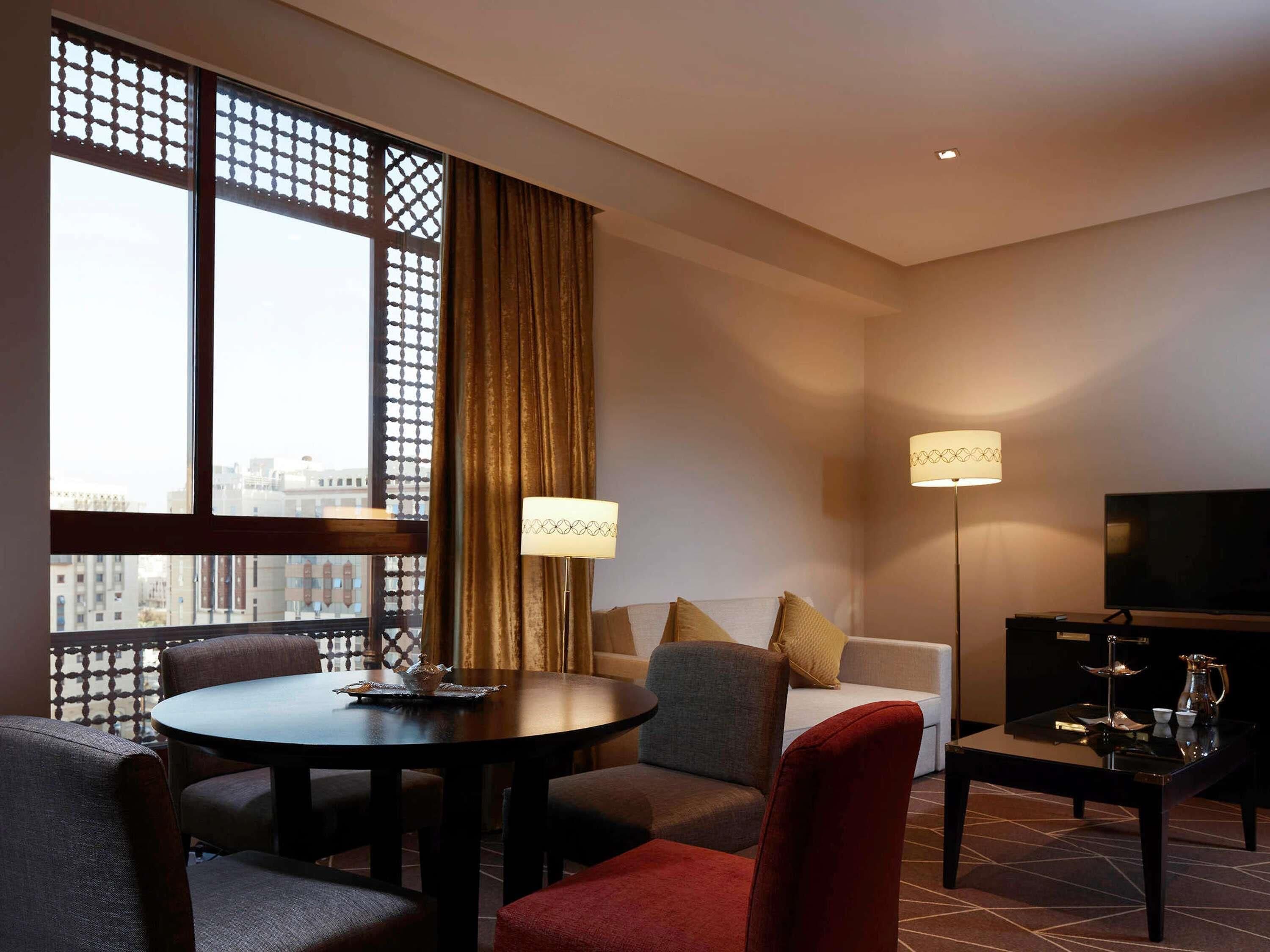 Habitación de hotel con mesa de comedor, sofá, televisor y vistas a la ciudad.
