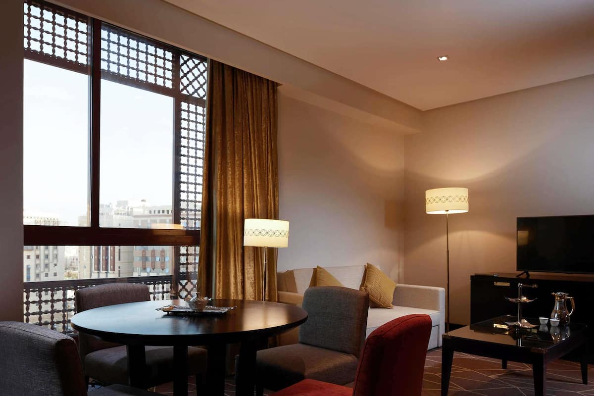 A hotel room with a dining table, sofa, TV, and a view of the city.
