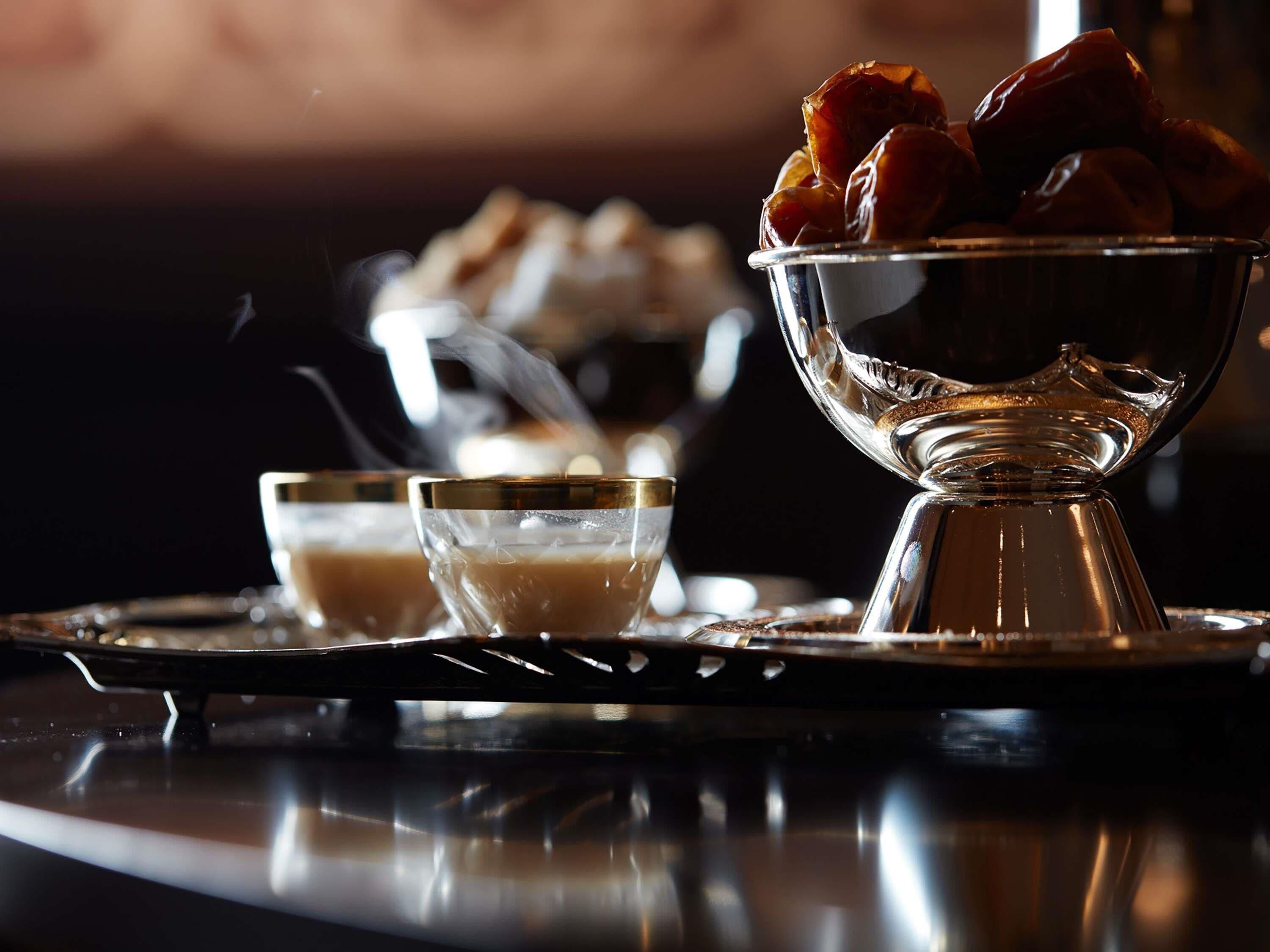 Un plateau avec un bol de dattes et deux verres contenant un liquide brun, probablement du thé ou du café, posés sur une surface réfléchissante.