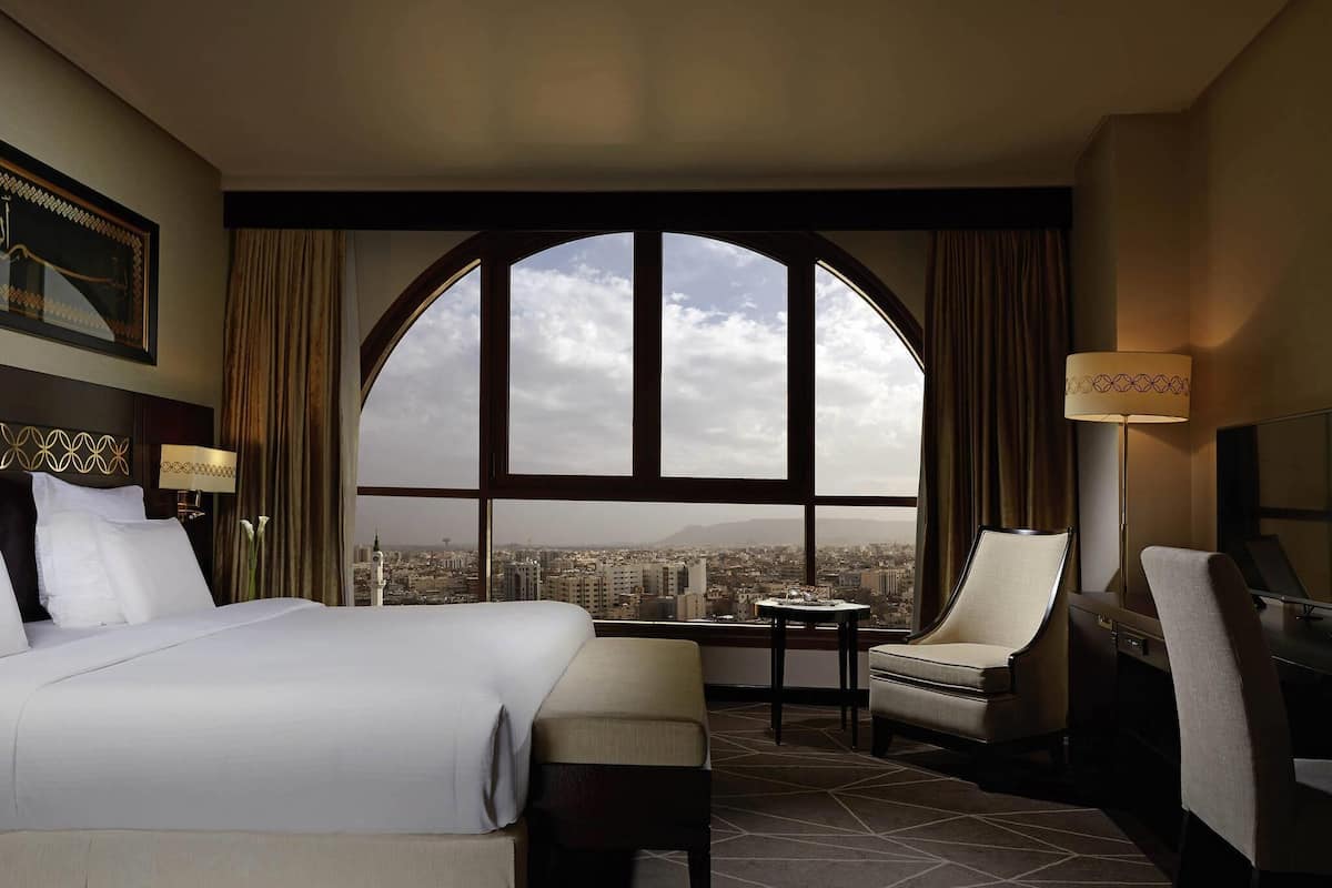 A hotel room with a large arched window, a bed, a desk, a chair, and a lamp.