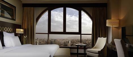 Una habitación de hotel con un gran ventanal arqueado, una cama, un escritorio, una silla y una lámpara.