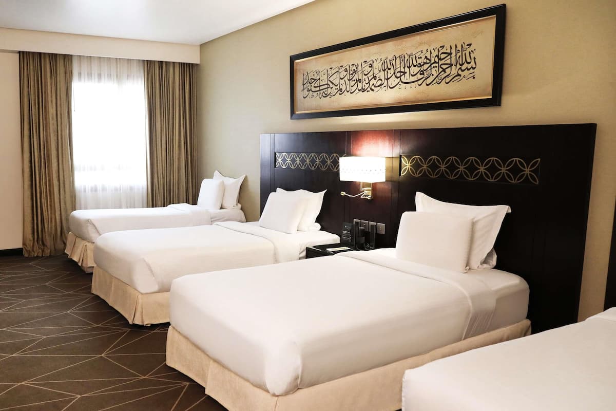 A hotel room with three beds, a large framed Arabic calligraphy piece, a modern light fixture, and a geometric patterned floor.