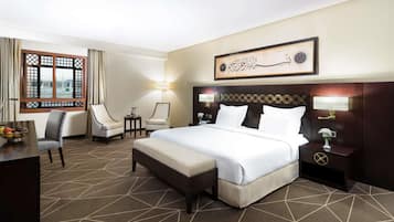 A hotel room with a large bed, two armchairs, a small table with a lamp, and a view of a mosque through a window.