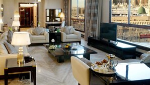 A spacious living room with a large TV, comfortable seating, a coffee table with fruits and pastries, and a view of a cityscape with a mosque.