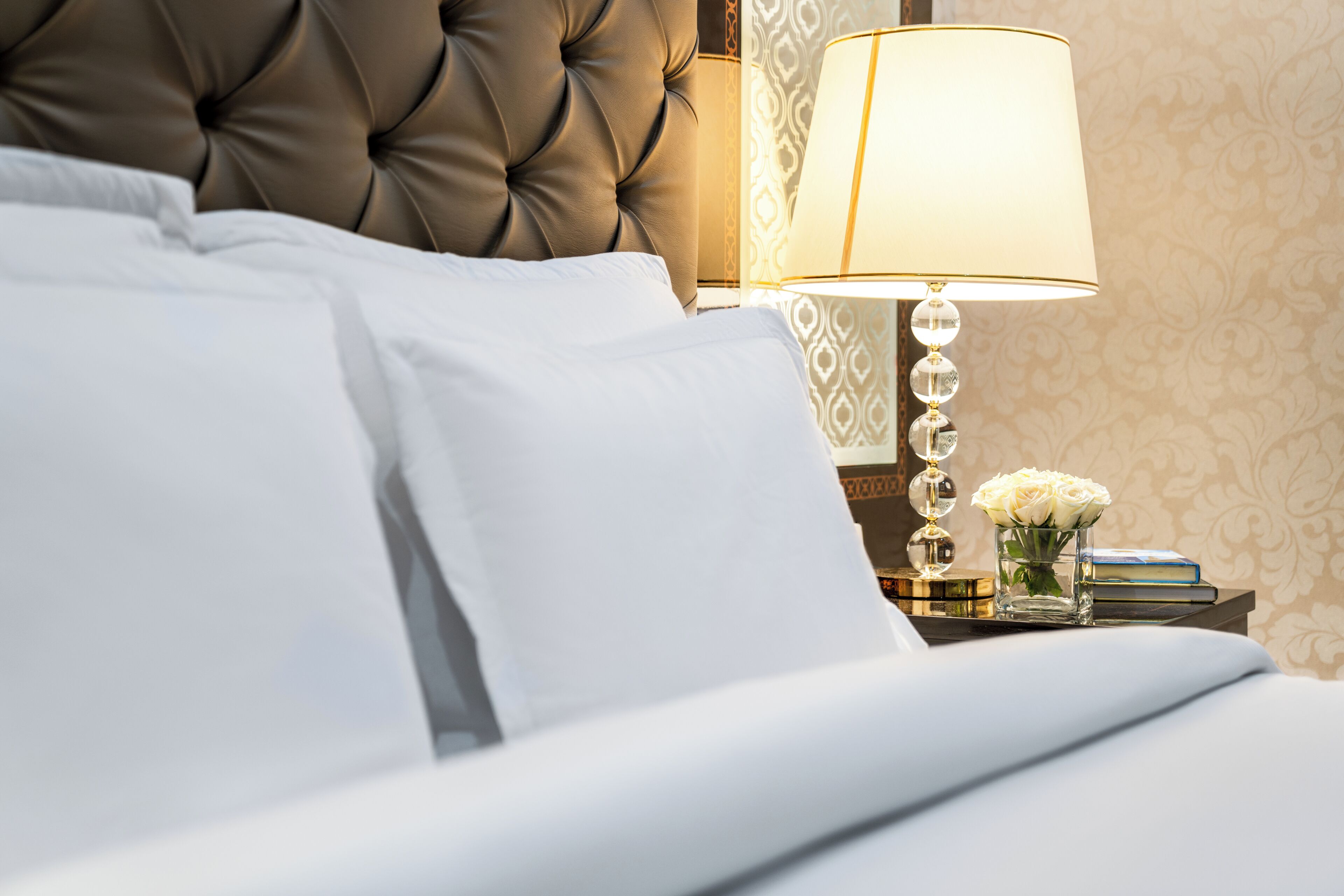 A neatly made bed with white linens, a tufted headboard, a bedside table with a lamp, and a vase with white flowers.