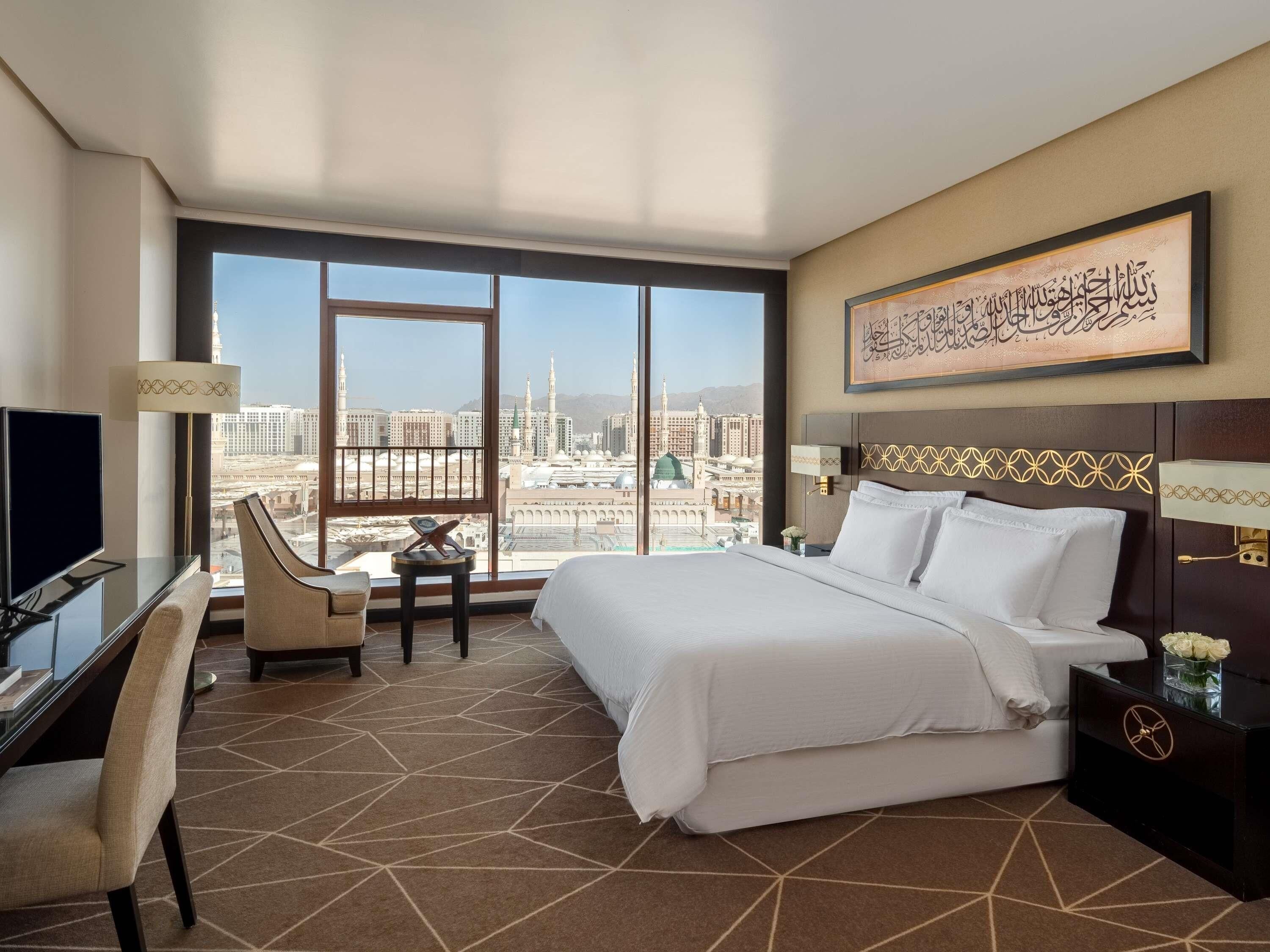 A hotel room with a large bed, a desk with a chair, a TV, and a view of a cityscape.