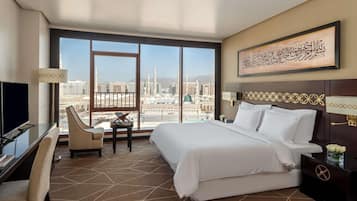 A hotel room with a large bed, a desk with a chair, a TV, and a view of a cityscape.